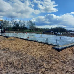 Świeżo wylana płyta fundamentowa z wystającymi prętami zbrojeniowymi i czarną folią kubełkową na obrzeżach, widoczny betonowóz w tle i niwelator laserowy na statywie.