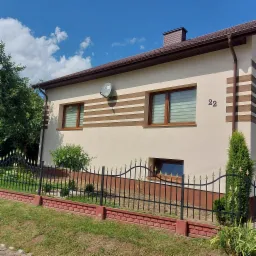 Elewacja domu jednorodzinnego w jasnym odcieniu z dekoracyjnymi, poziomymi pasami w kolorze brązowym, widoczne okna z roletami, numer domu '22' i antena satelitarna.