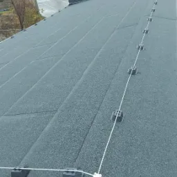 Bitumiczny dach płaski z zamontowanymi punktami kotwiczenia i liną asekuracyjną, widok z góry na tle jesiennego krajobrazu miejskiego.