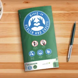 Broszura informacyjna o bezpieczeństwie i higienie pracy z logiem Heineken, leżąca na drewnianym stole obok miseczki pistacji i długopisu.