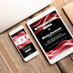 Tablet i smartfon wyświetlają reklamę wyprzedaży w salonie samochodowym Motor Centrum, leżące na drewnianym biurku obok koperty.