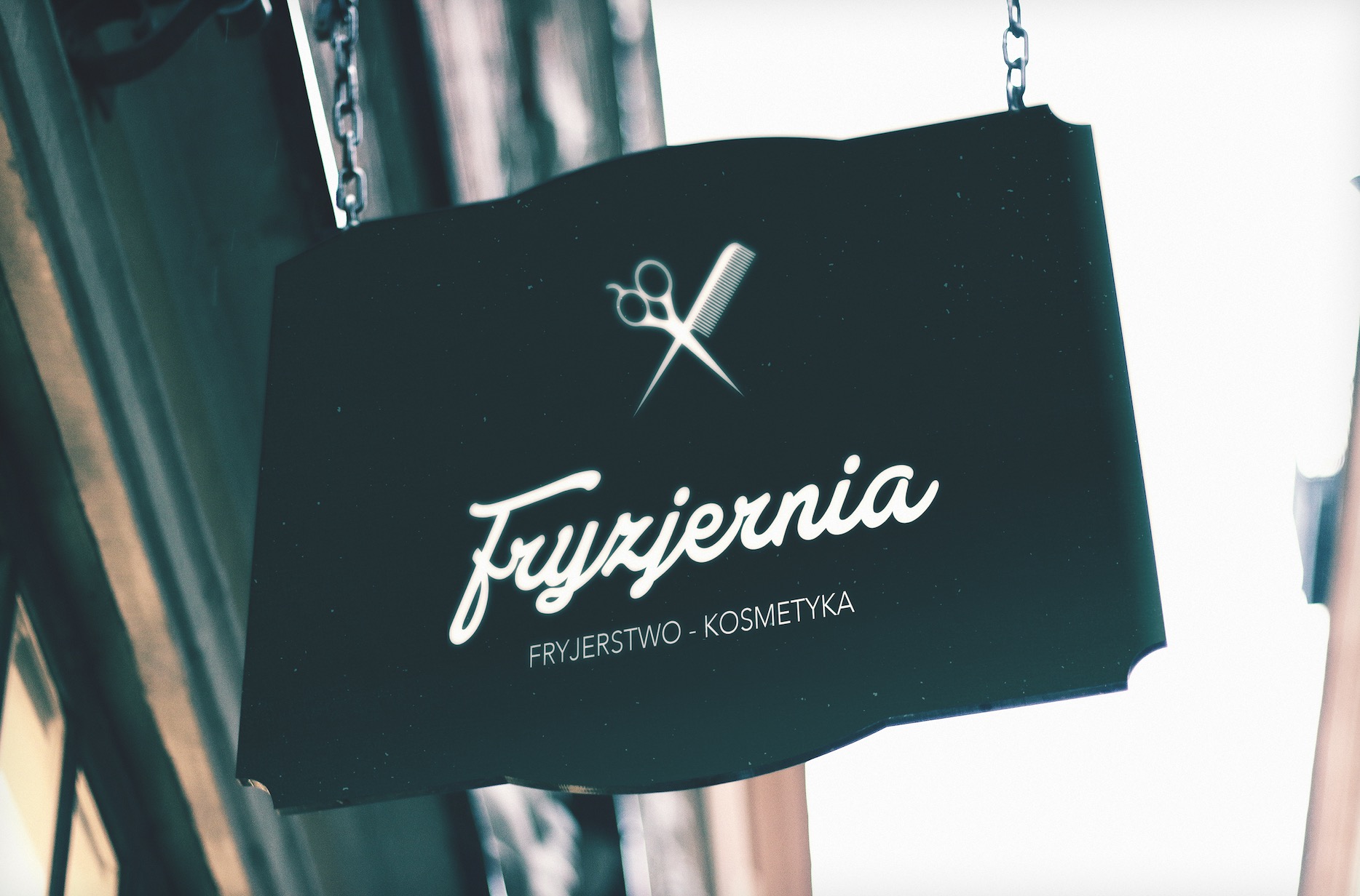 Czarny szyld wiszący na łańcuchu, prezentujący białe logo fryzjerskie z nożyczkami, grzebieniem i napisem 'Fryzjernia Fryzjerstwo-Kosmetyka' w ozdobnej czcionce.
