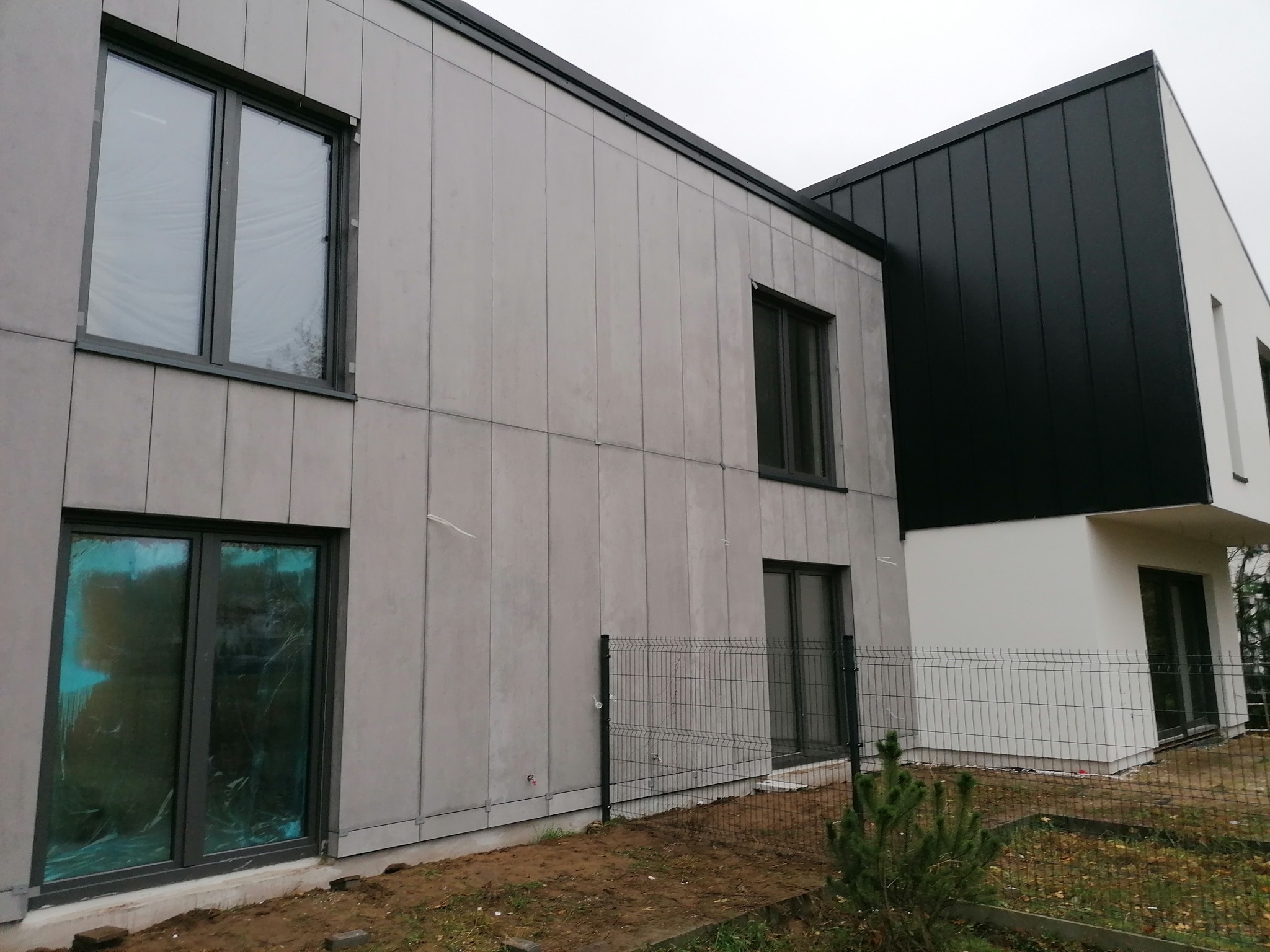 Nowoczesny dom z szarą elewacją panelową i czarnym dachem, jedno okno zabezpieczone folią ochronną, widok z zewnątrz na ogrodzenie i fragment ogrodu.