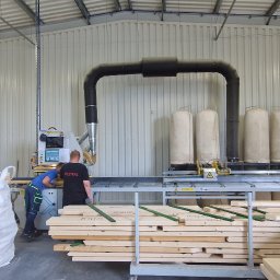 MISTRAL KWIATKOWSKI I WSPÓLNICY SPÓŁKA KOMANDYTOWA - Praca przy obróbce drewna w hali: pracownicy obsługują maszynę do cięcia, w tle filtry powietrza, na pierwszym planie ułożone deski i worek z odpadami.