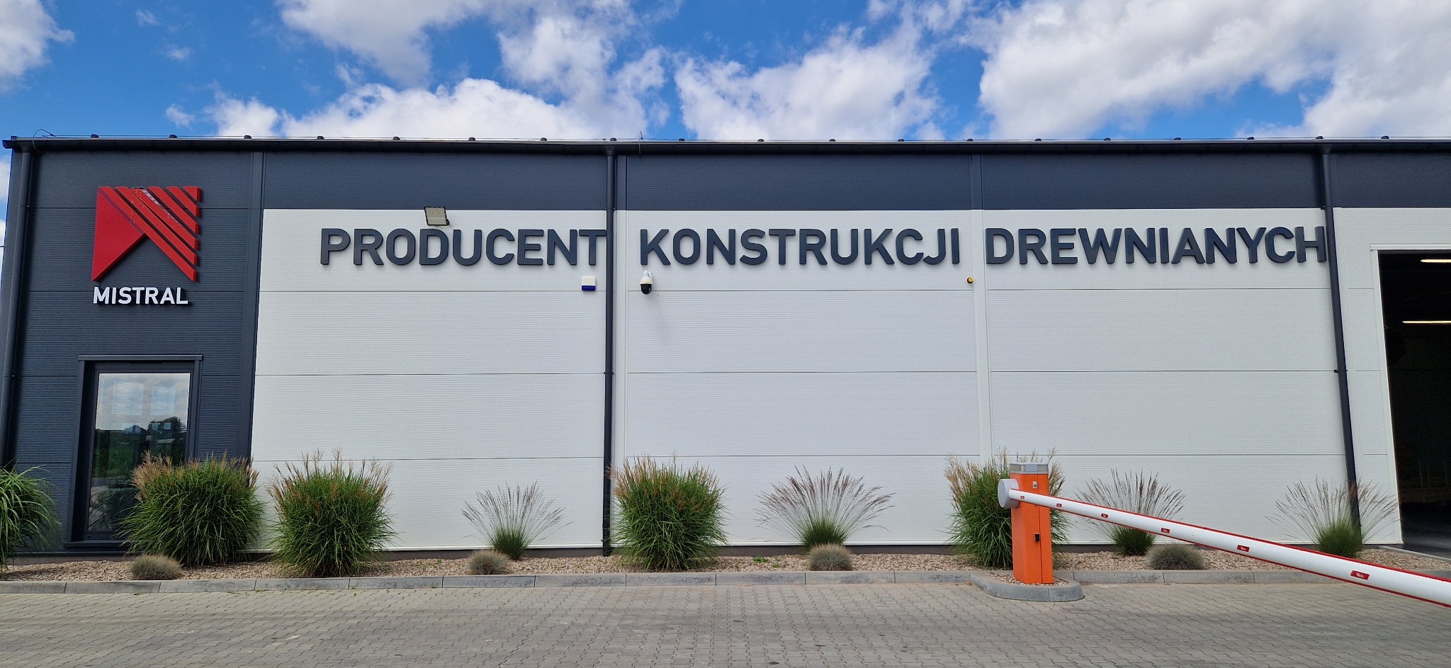 Budynek firmy Mistral, producenta konstrukcji drewnianych, z logo i napisem na elewacji. Widoczne krzewy ozdobne i szlaban wjazdowy. Błękitne niebo z chmurami w tle.