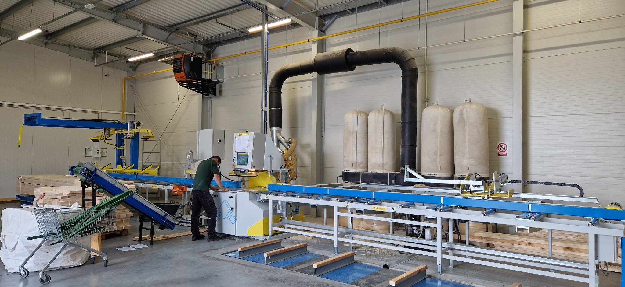 Pracownik obsługuje maszynę do obróbki drewna w hali produkcyjnej. Widoczne elementy linii produkcyjnej, filtry powietrza i składowane drewno.