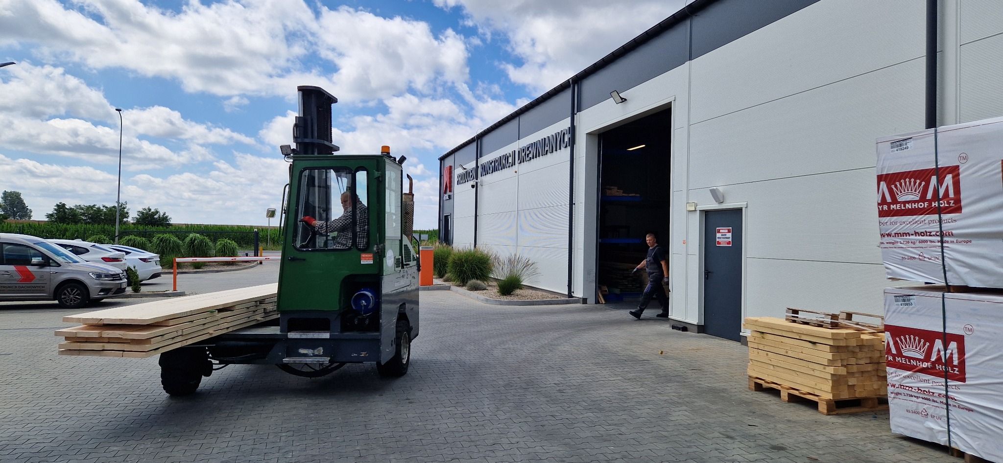 Wózek widłowy z ładunkiem drewna na tle magazynu z napisem 'Producent konstrukcji drewnianych'. Pracownik wchodzi do magazynu. Składowane paczki drewna.