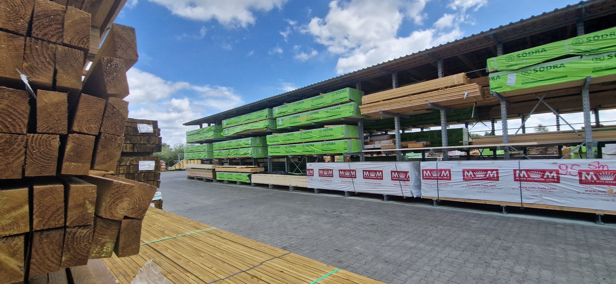 Składowane drewno budowlane na regałach magazynowych, widoczne pakiety z logo producentów, błękitne niebo w tle. Drewno konstrukcyjne.