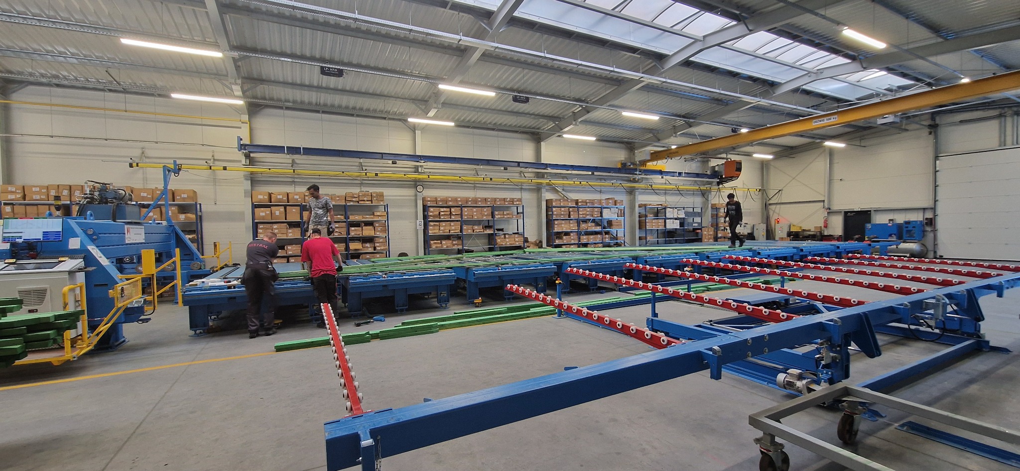 Proces obróbki drewna w hali produkcyjnej: pracownicy obsługują maszynę do cięcia, w tle regały z kartonami. Widoczny surowiec i elementy konstrukcyjne.