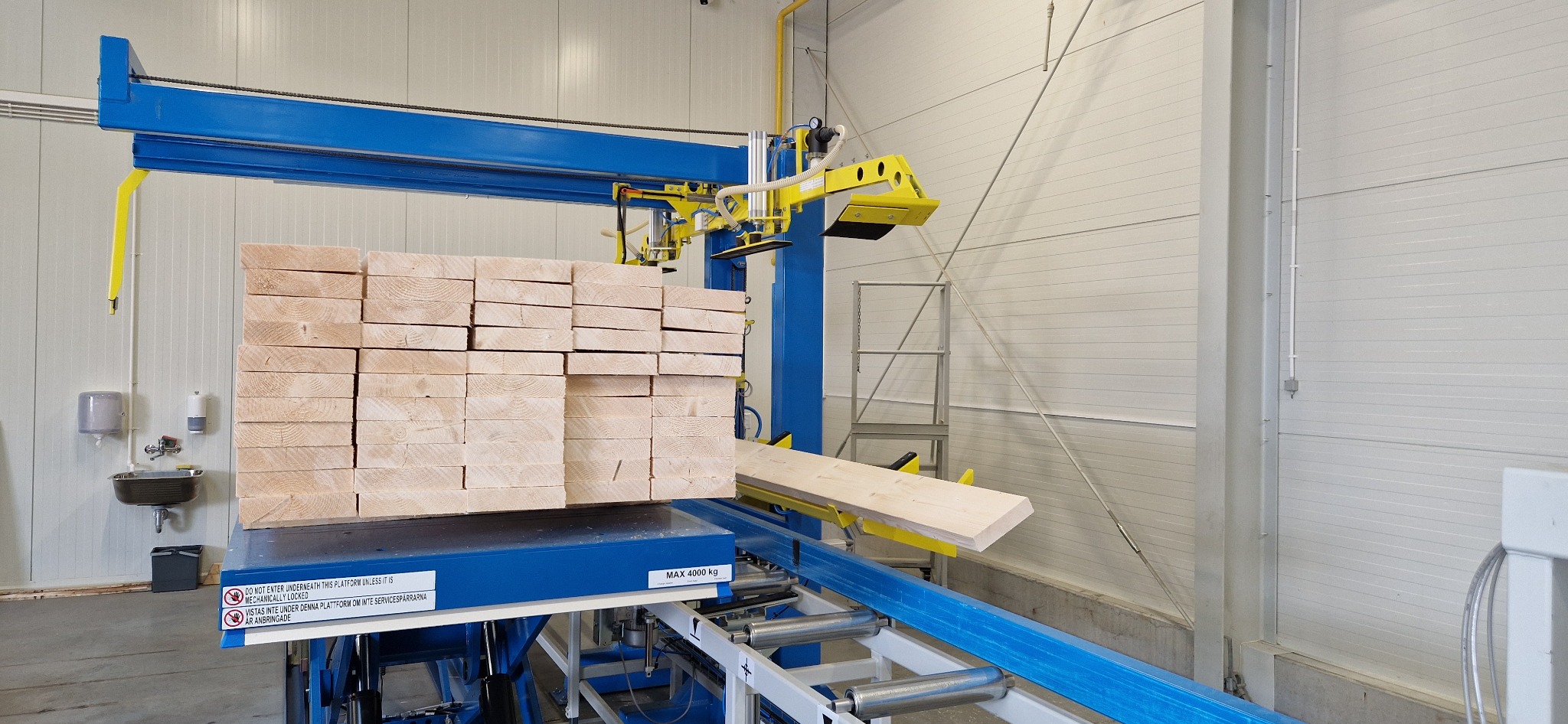 Skład drewna: automatyczny podajnik desek z niebiesko-żółtą konstrukcją, ułożone deski na platformie. Widoczne oznaczenia bezpieczeństwa i maksymalne obciążenie.