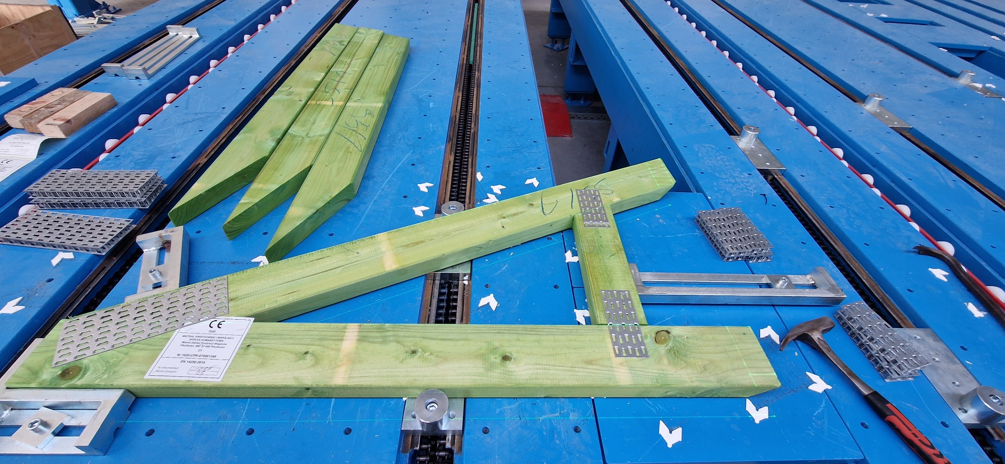Elementy więźby dachowej z łącznikami na niebieskim stole montażowym. Widoczne narzędzia i etykieta CE. Drewno impregnowane na zielono.