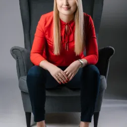 Portret kobiety siedzącej w fotelu typu uszak, ubrana w czerwoną bluzkę i granatowe spodnie, z zegarkiem na ręce i pierścionkiem, uśmiechnięta.