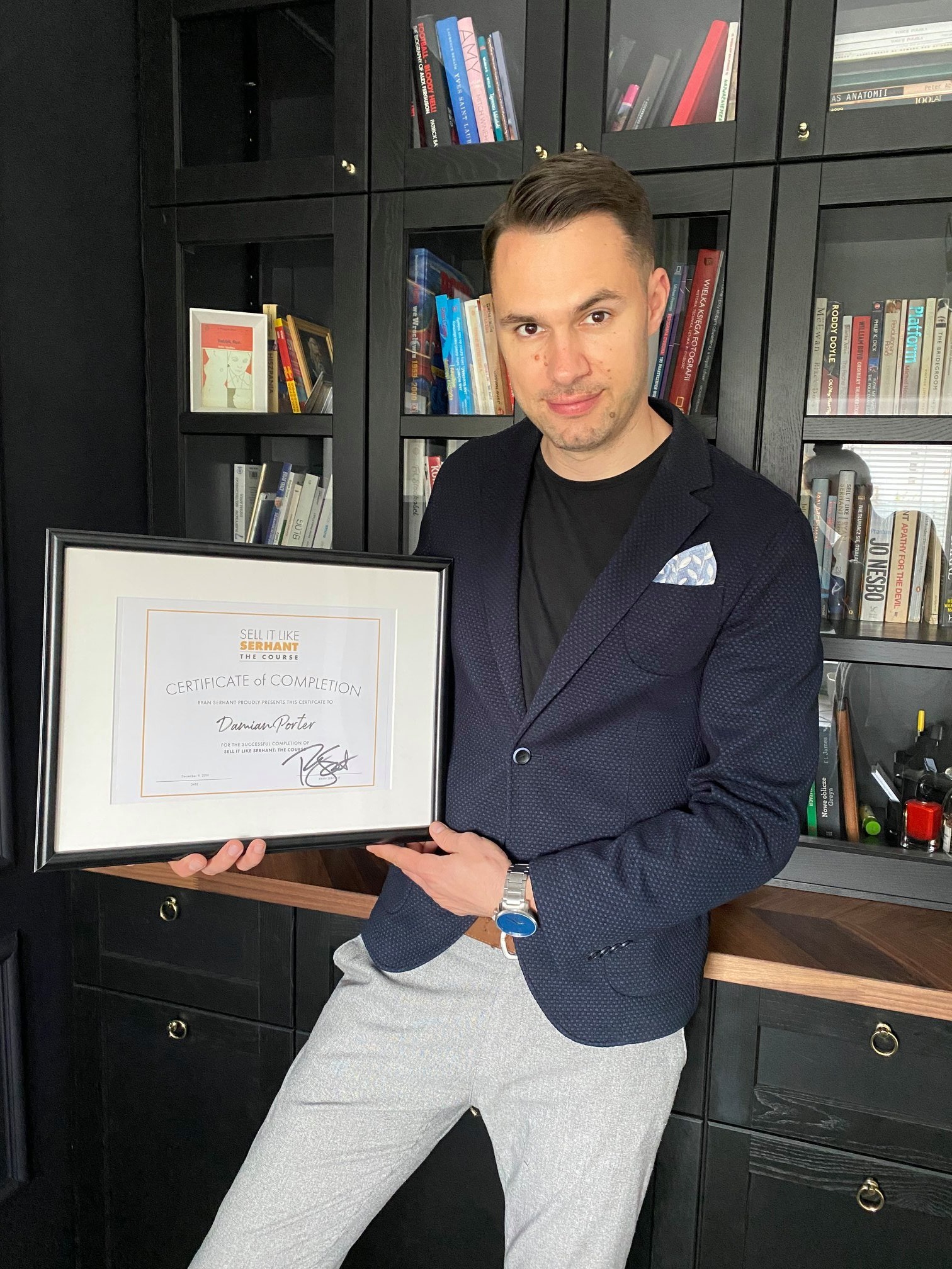Mężczyzna w marynarce prezentuje certyfikat ukończenia kursu 'Sell It Like Serhant' na tle regału z książkami.