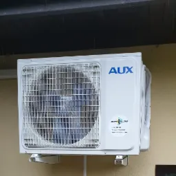 Zewnętrzna jednostka klimatyzacji AUX zamontowana na ścianie budynku, widoczne logo firmy Monk Klima na urządzeniu, lekki deszcz.