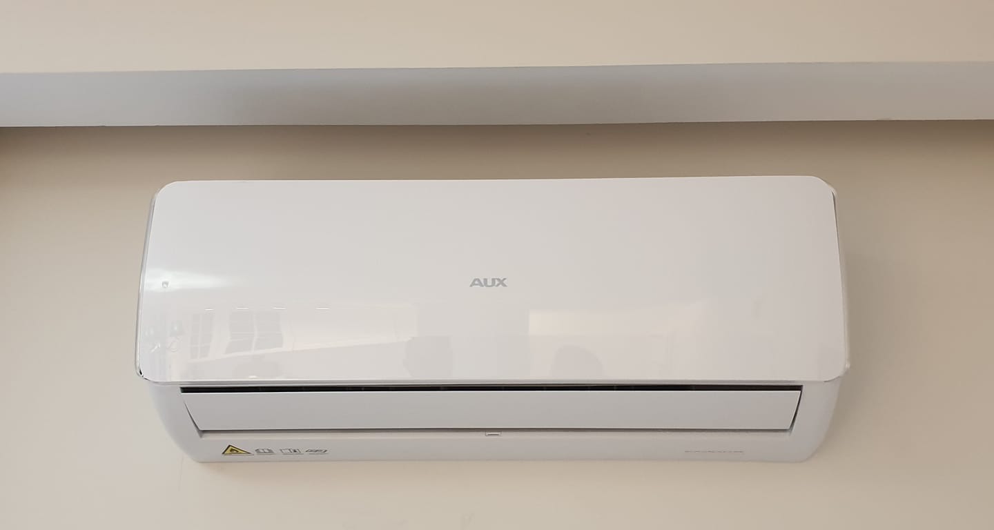 Biała klimatyzacja ścienna marki AUX zamontowana na beżowej ścianie, widoczne oznaczenia bezpieczeństwa na obudowie.