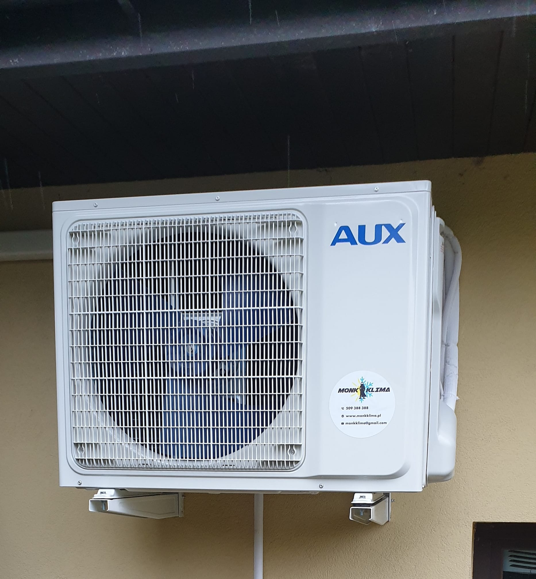 Zewnętrzna jednostka klimatyzacji AUX zamontowana na ścianie budynku, widoczne logo firmy Monk Klima na urządzeniu, lekki deszcz.