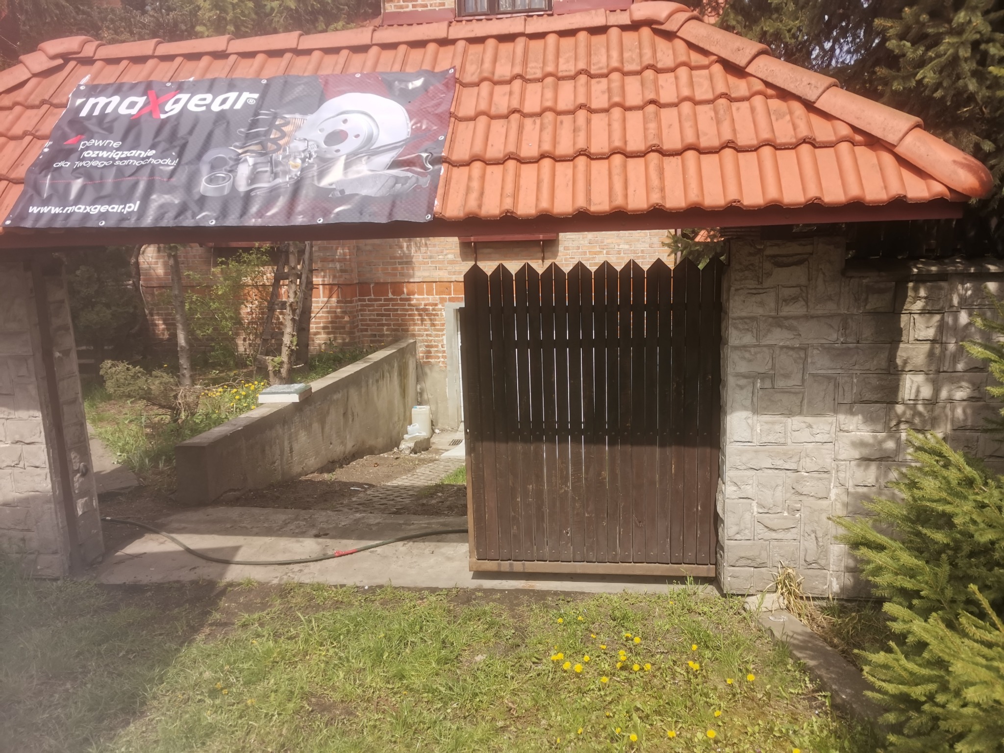 Wjazd na posesję z drewnianą bramą i banerem reklamowym 'maxgear' z grafiką części samochodowych pod zadaszeniem z czerwonej dachówki.