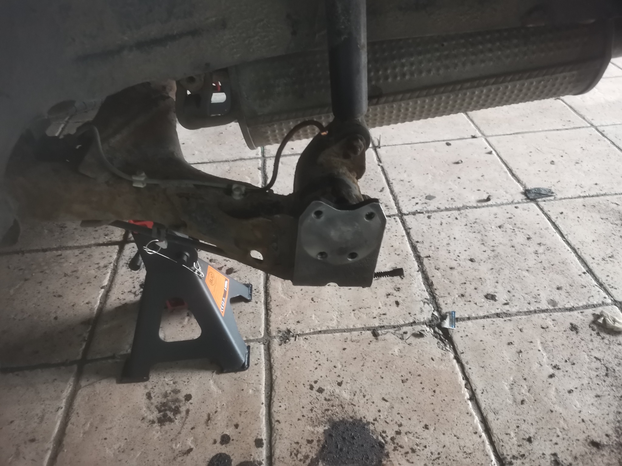 Zawieszenie samochodu podparte na podnośniku warsztatowym, widoczne elementy układu jezdnego i wydechowego, zabrudzona podłoga warsztatu z plamami oleju.