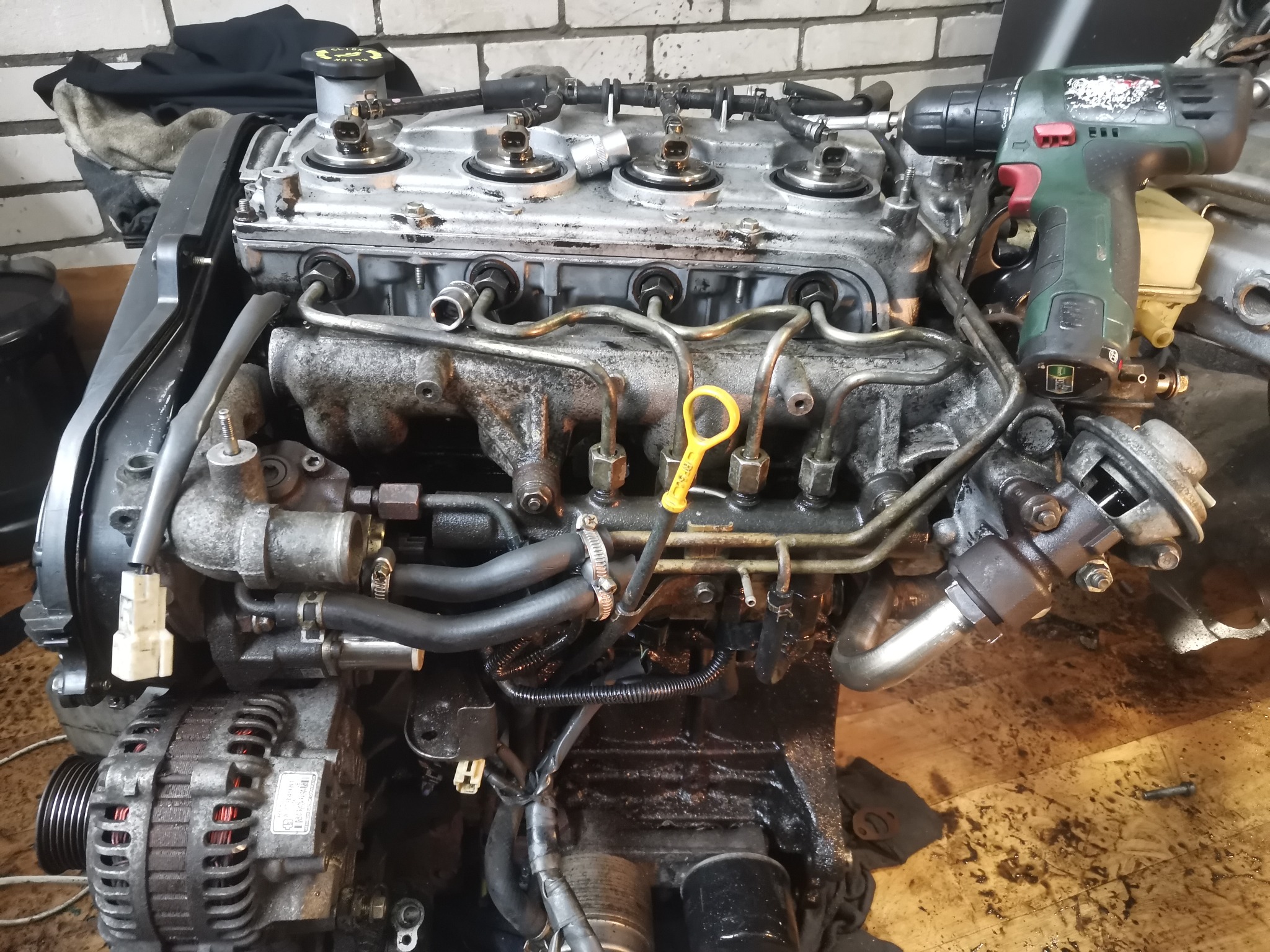 Silnik samochodowy z widocznymi przewodami paliwowymi, alternatorem i wkrętarką akumulatorową na tle ceglanej ściany.