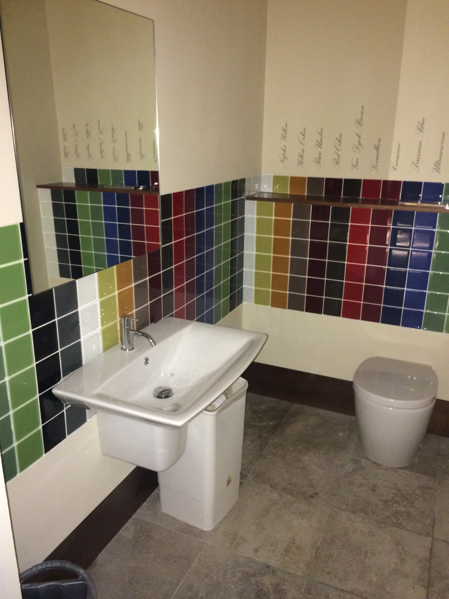 Łazienka z umywalką na postumencie i toaletą, ściany wyłożone kolorowymi kwadratowymi płytkami, nad którymi widnieją nazwy kolorów farb.