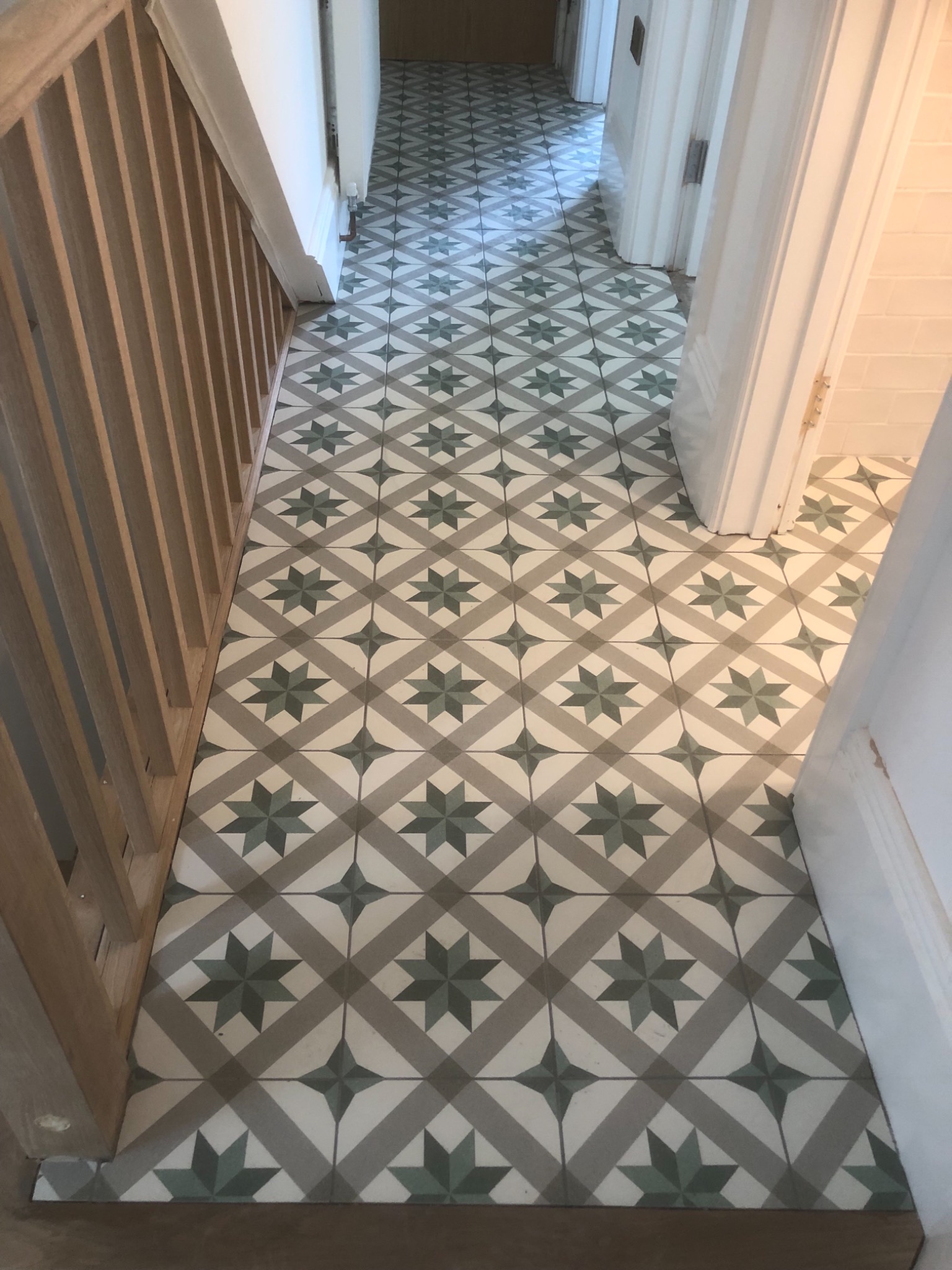 Wzorzysta podłoga z płytek ceramicznych w odcieniach szarości i zieleni, ułożona w korytarzu, widoczna przy drewnianej balustradzie schodów i otwartych drzwiach do innego pomieszczenia.