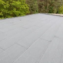 Nowo pokryty, płaski dach z papy termozgrzewalnej w kolorze szarym, widoczne łączenia, w tle zieleń drzew, na dachu wiaderko z wodą.