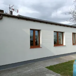 Boczny widok świeżo otynkowanego domu z brązowymi oknami i rynnami, widoczna antena na dachu i niebieska beczka na trawniku.