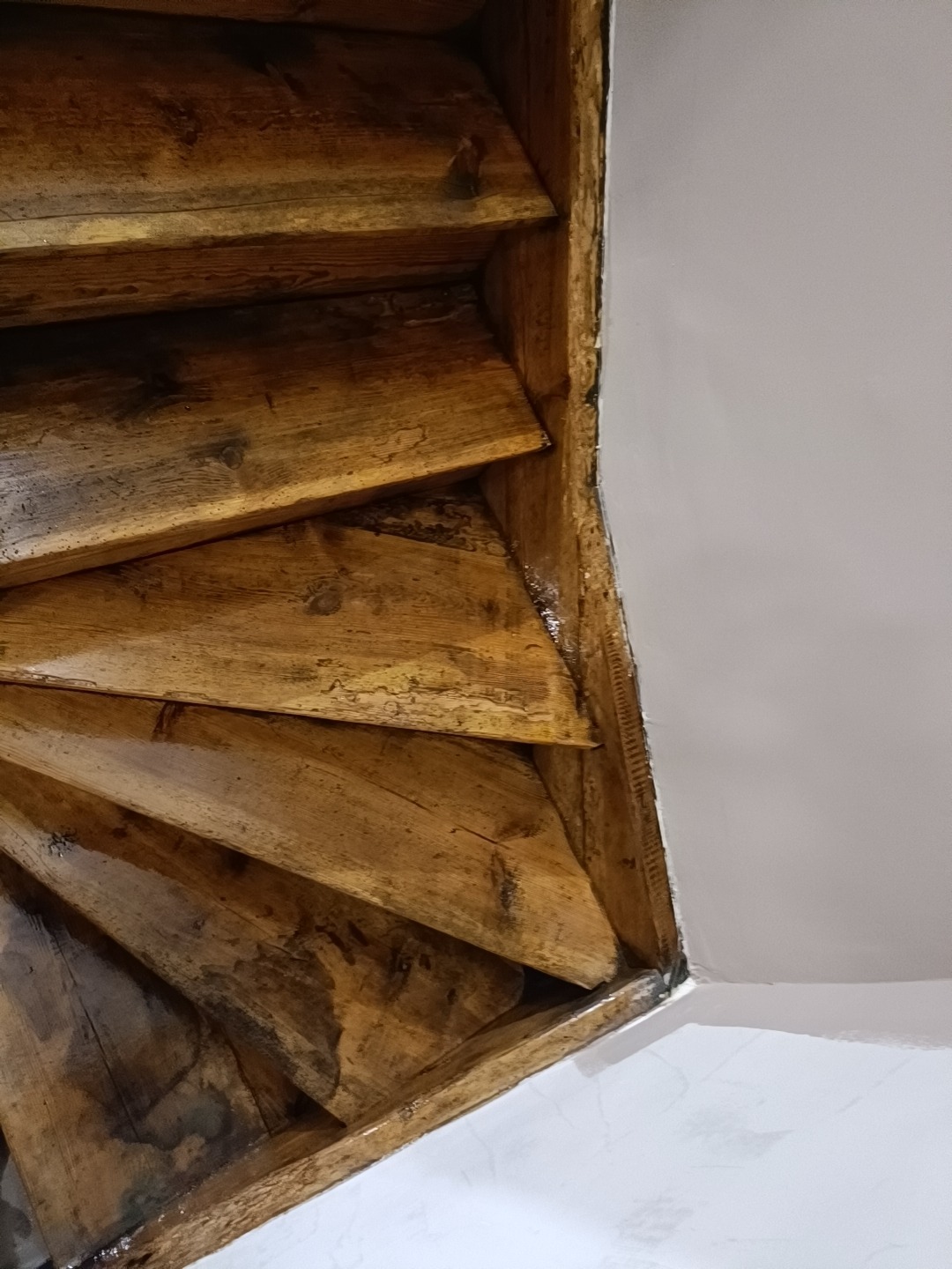 Drewniane schody spiralne, widok z dołu, ukazujący detale konstrukcji i połączenie z białą ścianą.