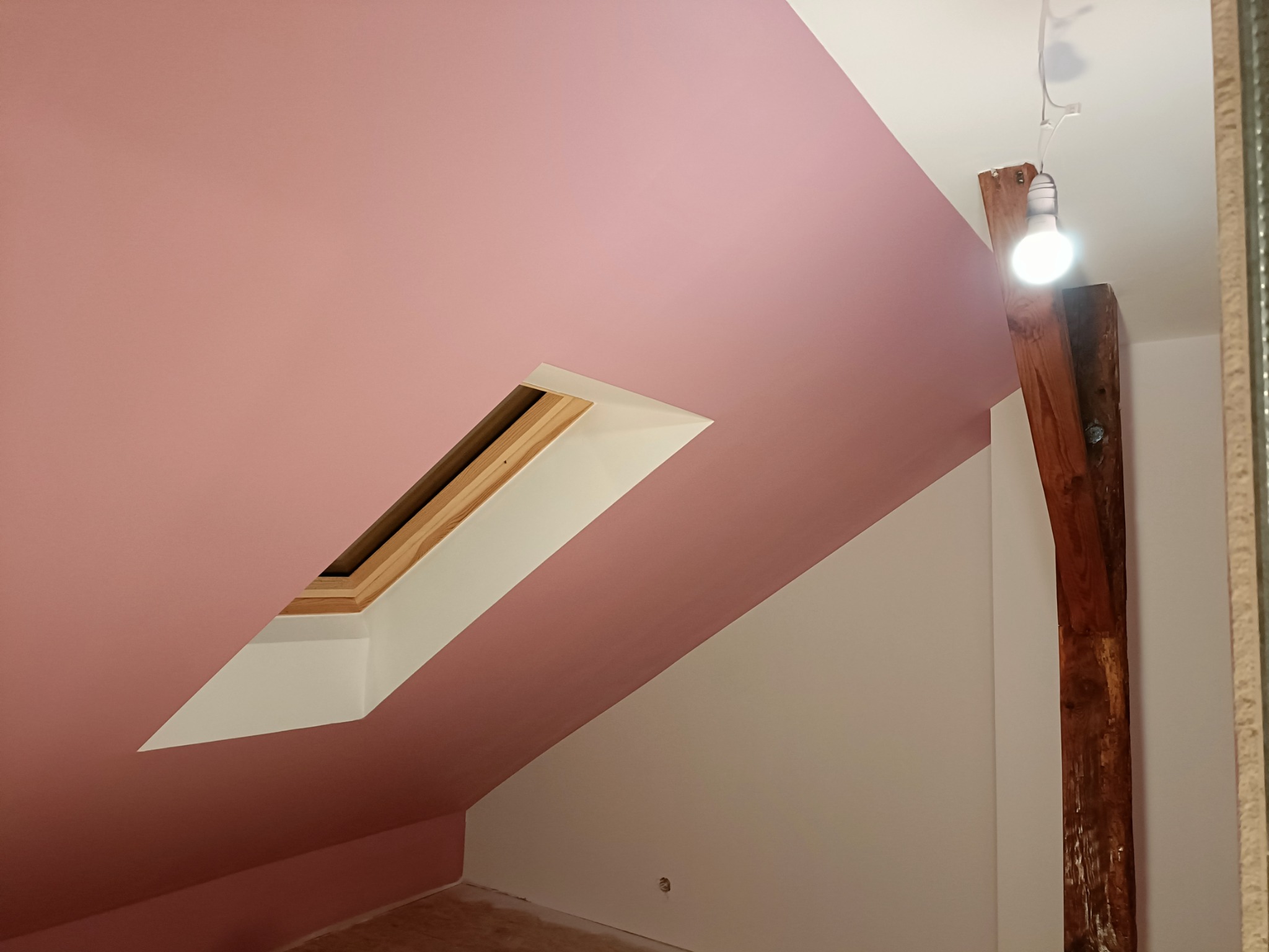 Poddasze po malowaniu na różowo, widoczne okno dachowe z drewnianą ramą, surowy drewniany słup z żarówką i fragment ściany pomalowanej na biało.