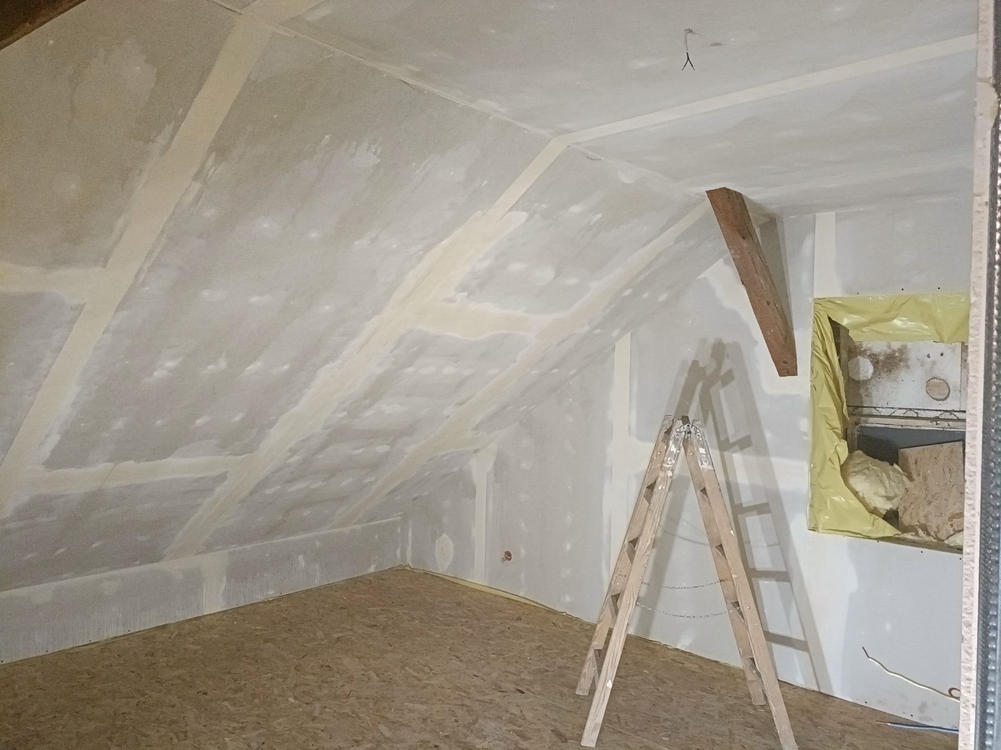 Poddasze w trakcie wykończenia: płyty gipsowo-kartonowe z widocznymi łączeniami, drewniana drabina oparta o ścianę, izolacja zabezpieczona folią w otworze okiennym, surowe drewno na skosie dachu.