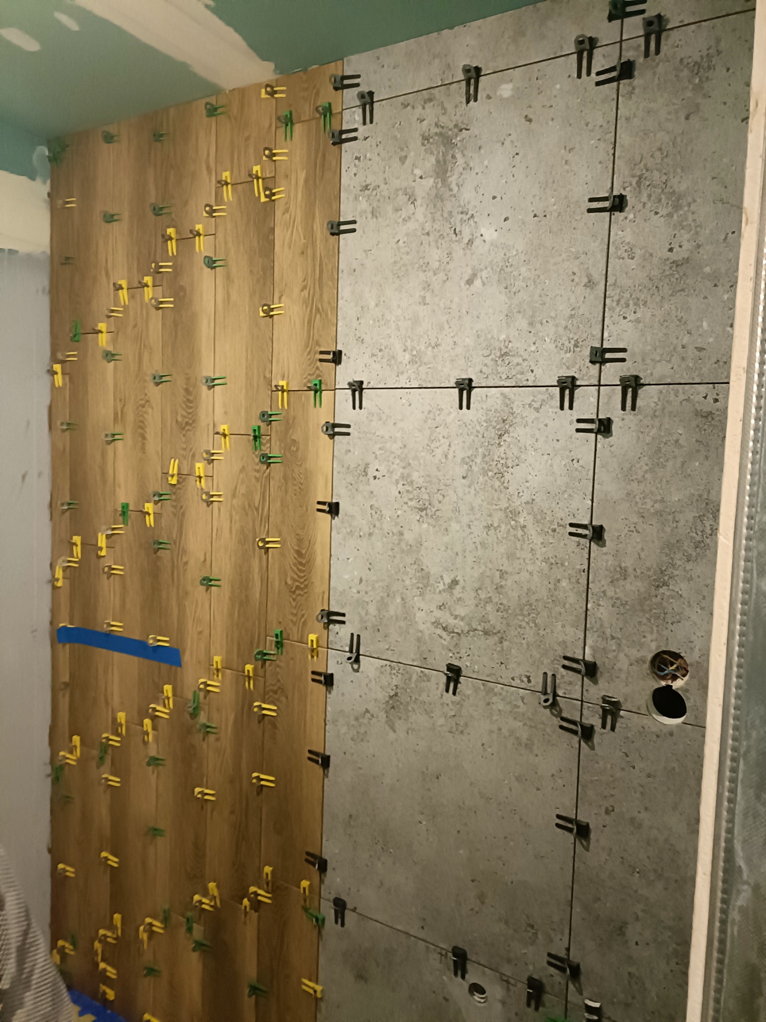 Położone do połowy płytki imitujące drewno i płytki betonowe na ścianie, z użyciem systemu poziomowania płytek z żółtymi i zielonymi klinami oraz czarnymi klipsami.