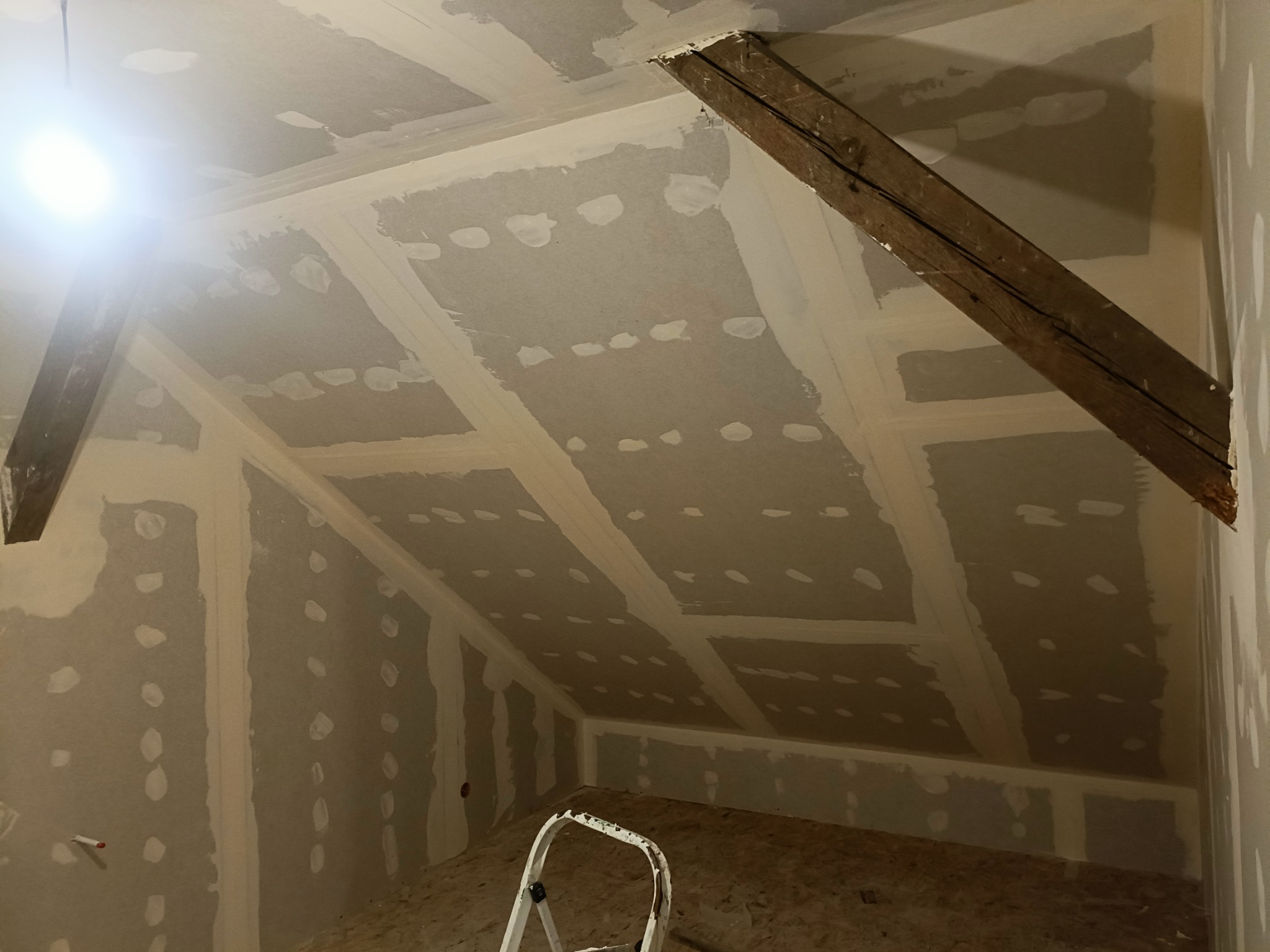 Poddasze w trakcie wykańczania: płyty gipsowo-kartonowe z widocznymi łączeniami i szpachlowanymi wkrętami, drewniana belka konstrukcyjna, drabina w pomieszczeniu.
