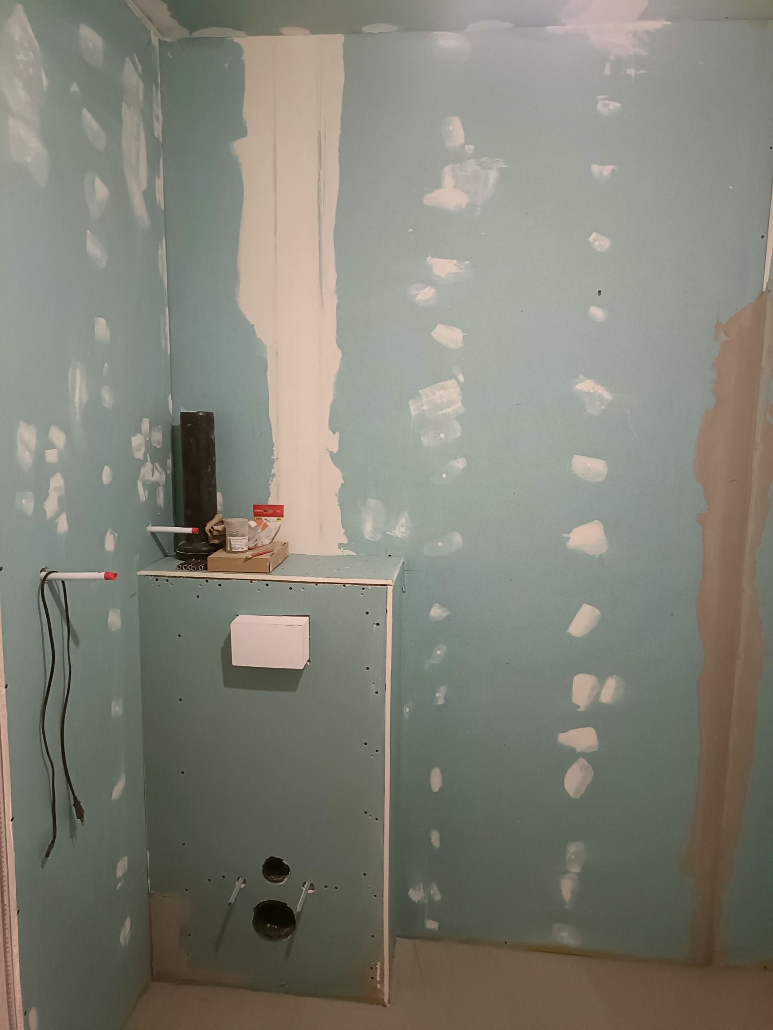 Wnętrze łazienki w trakcie prac wykończeniowych: ściany z płyt gipsowo-kartonowych z widocznymi punktami szpachlowania, przygotowane pod instalację sanitarną, z widocznymi rurami i akcesoriami.