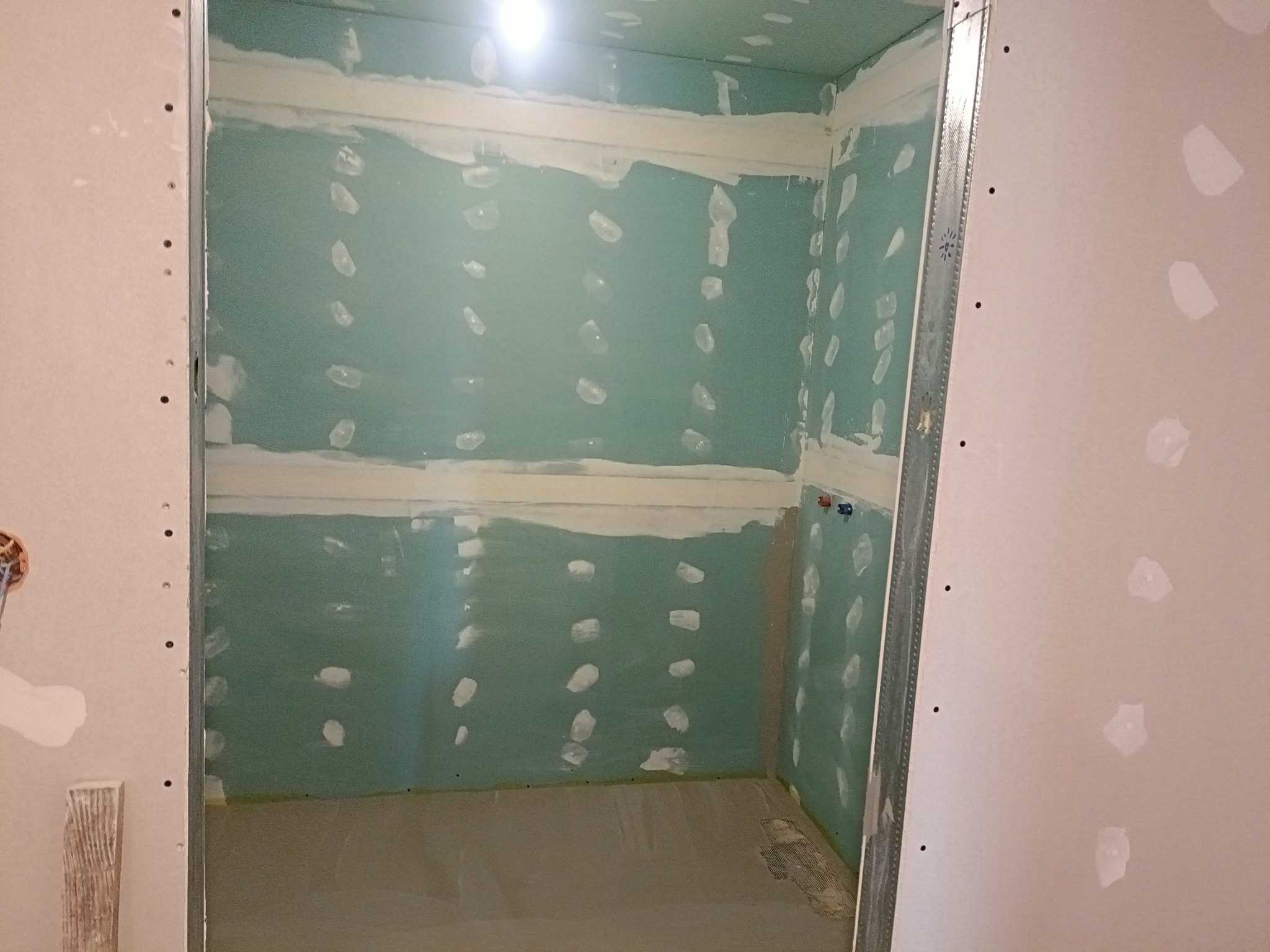 Wnętrze pomieszczenia w trakcie prac wykończeniowych: płyty gipsowo-kartonowe na ścianach i suficie, widoczne łączenia płyt zaszpachlowane, metalowe profile konstrukcyjne, podłoga zabezpieczona folią.