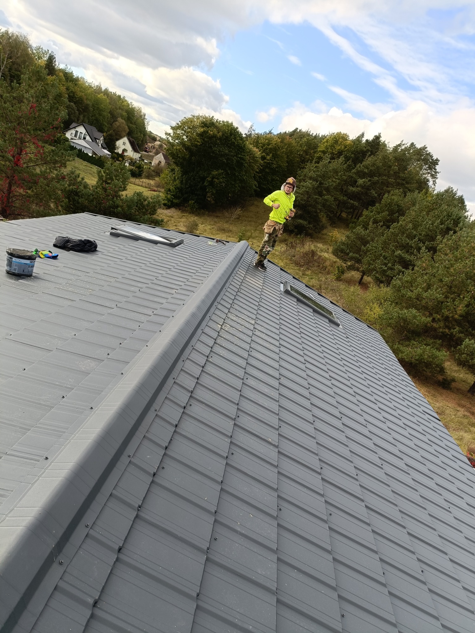 Pracownik w jaskrawej kurtce stoi na nowym, szarym dachu z blachy trapezowej, z widocznymi oknami dachowymi i narzędziami na krawędzi, na tle zieleni drzew i błękitnego nieba z chmurami.