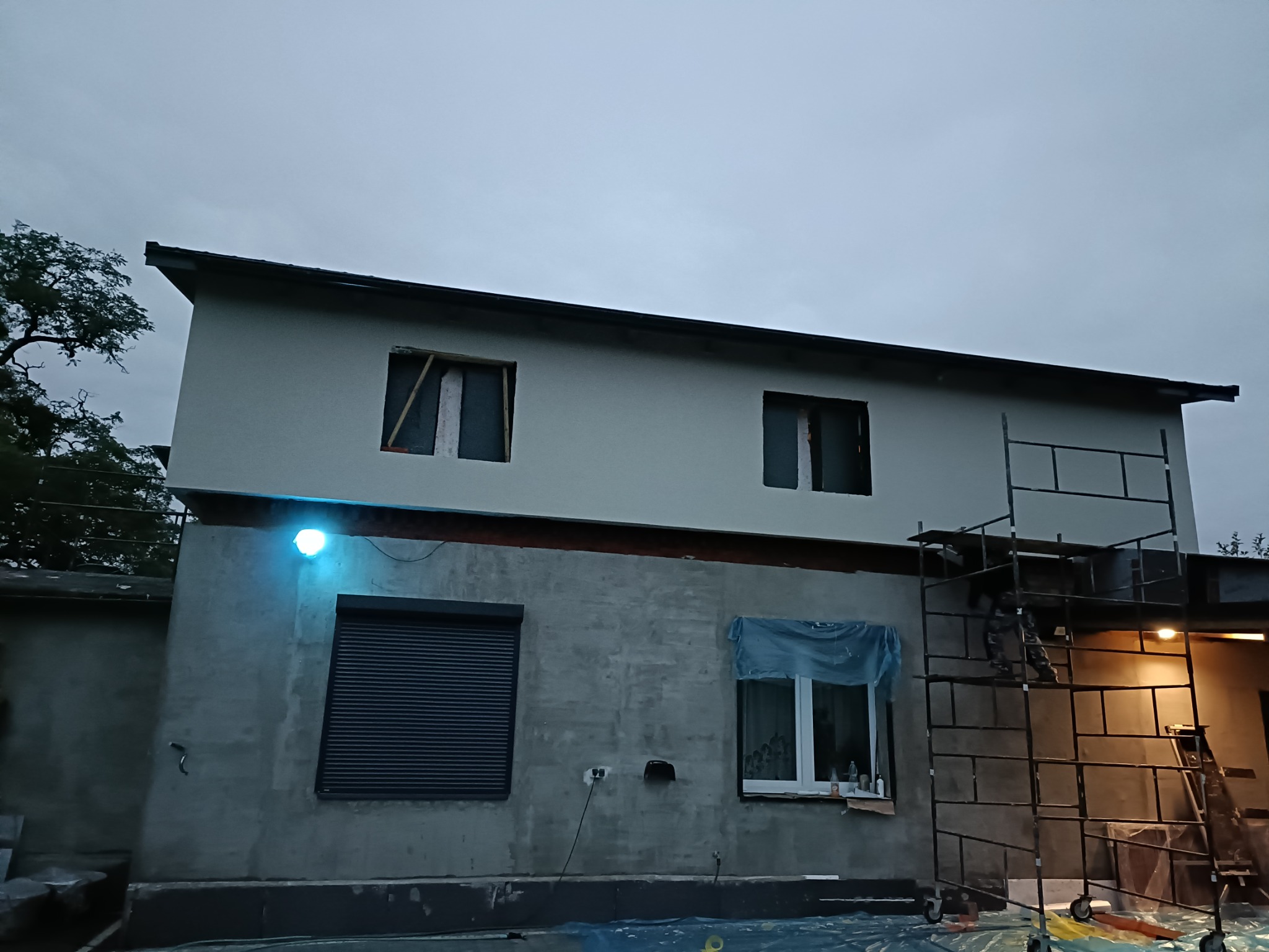 Dwukondygnacyjny budynek w trakcie prac elewacyjnych: otynkowana górna część i surowa, szara dolna, z widocznymi rusztowaniami i oświetleniem budowlanym, uchwycony przy pochmurnym niebie.