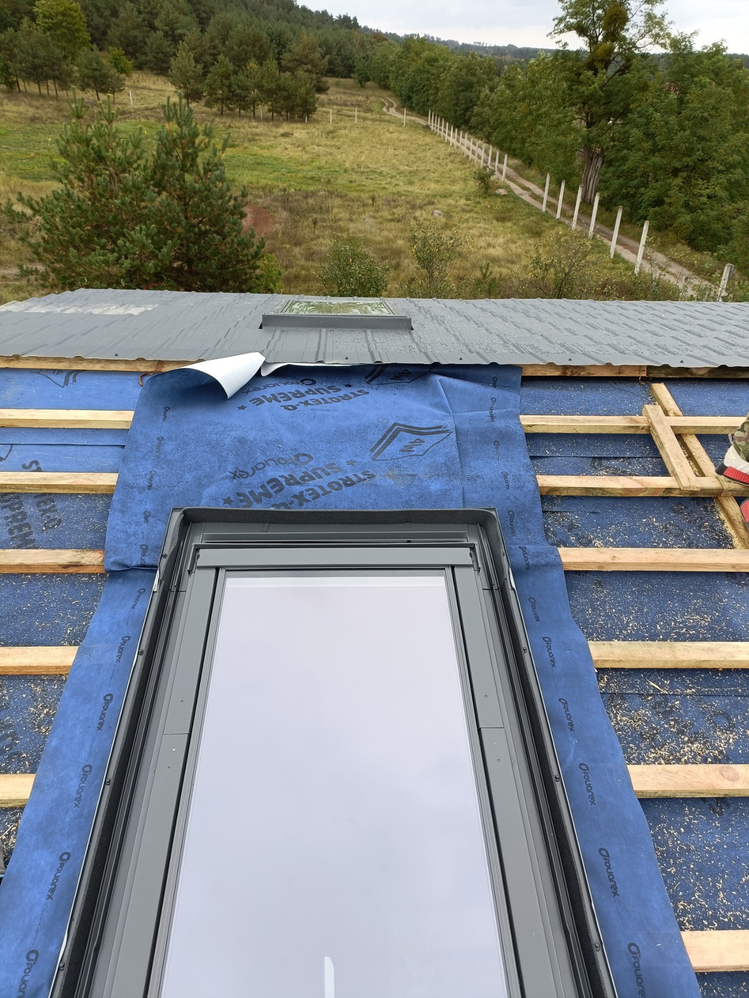 Montaż okna dachowego: widok z góry na wstawione okno połaciowe, zabezpieczone niebieską membraną dachową Stroetex Supreme, z widocznymi łatami i fragmentem pokrycia dachowego z blachy trapezowej...