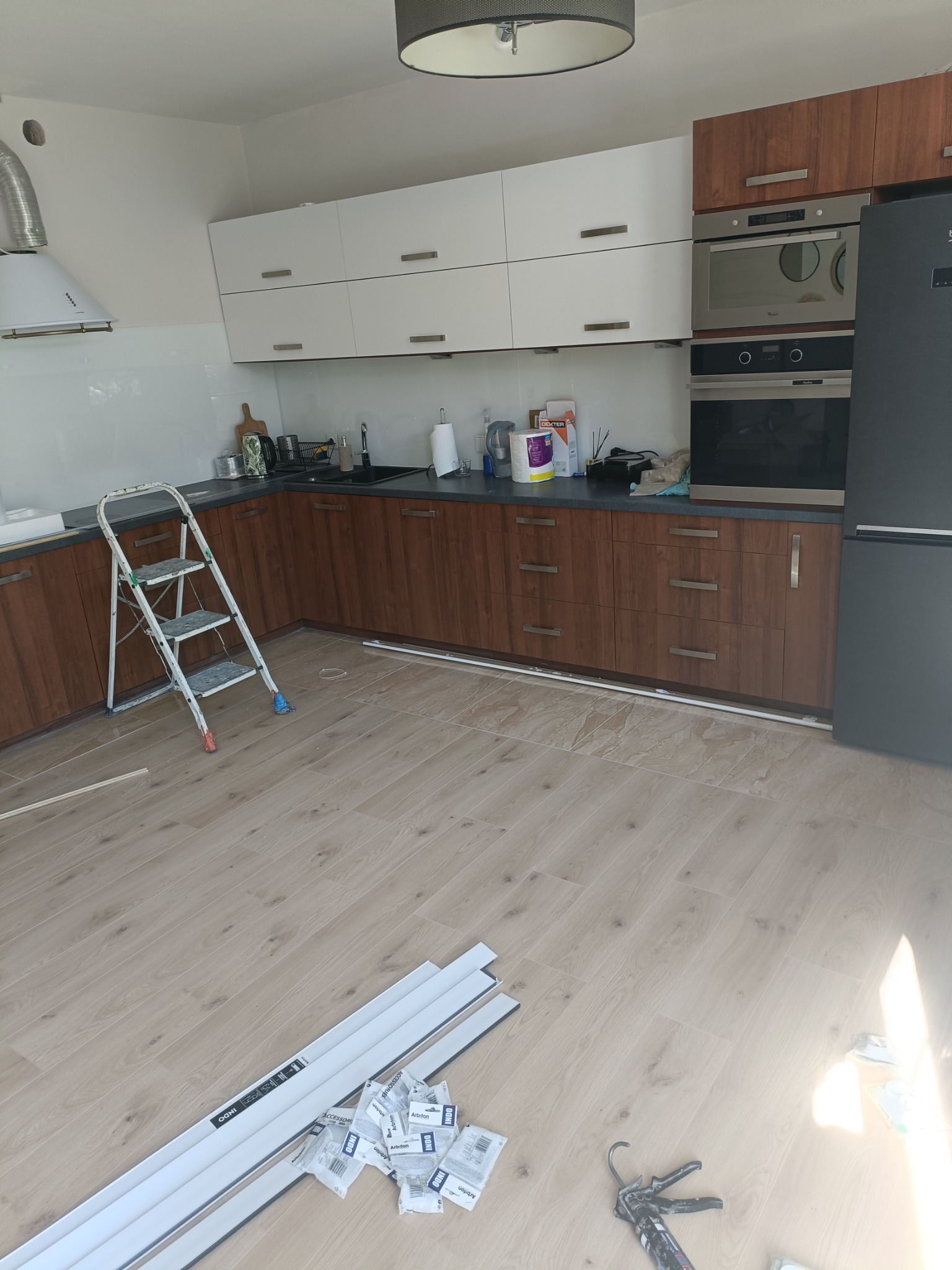 Wnętrze kuchni w trakcie montażu listew przypodłogowych; widoczna drabina, białe i brązowe szafki, sprzęt AGD oraz akcesoria montażowe na podłodze.