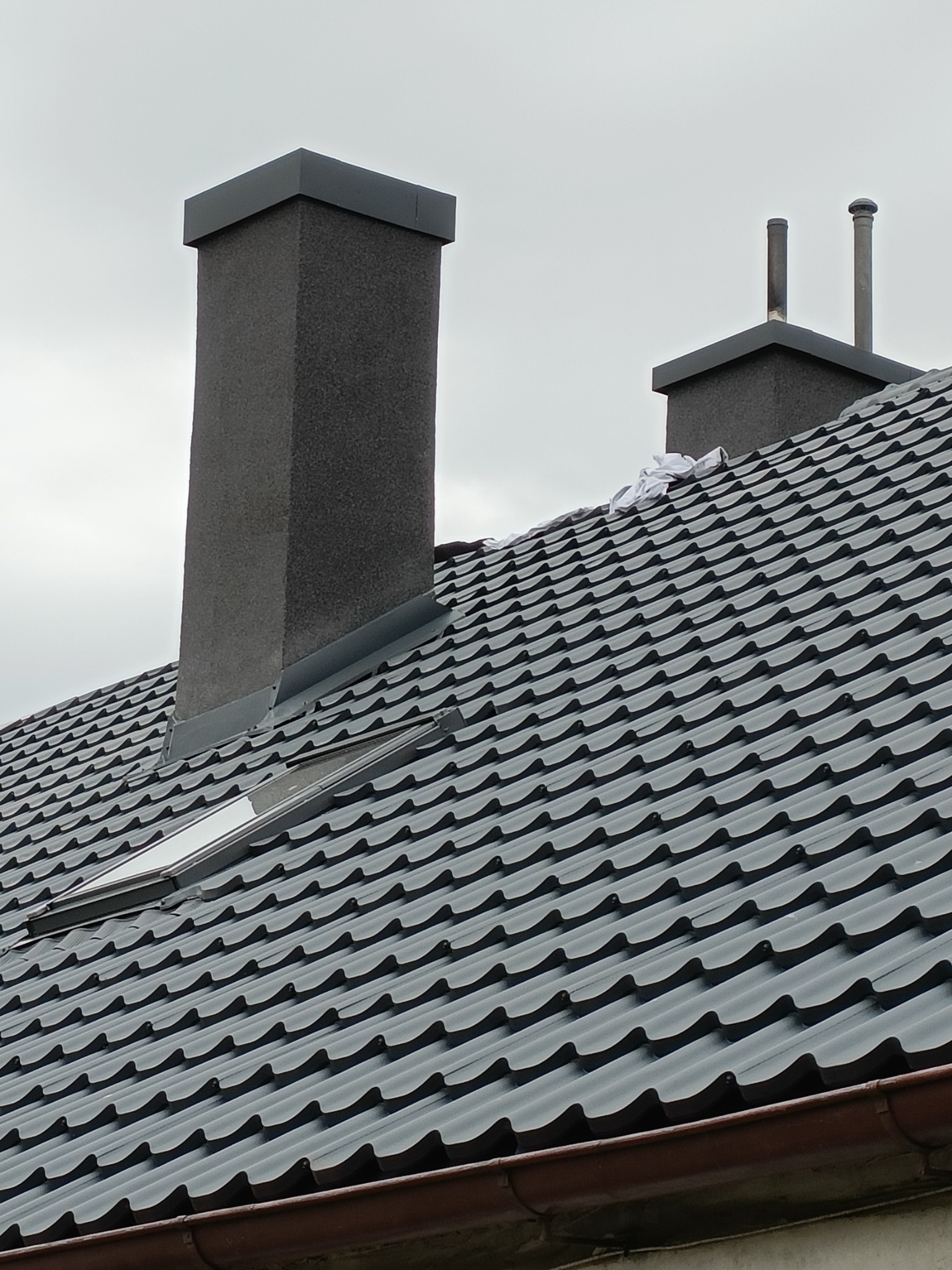 Grafitowy dach z blachodachówki z dwoma kominami o różnej wysokości i oknem dachowym, widok z dołu.