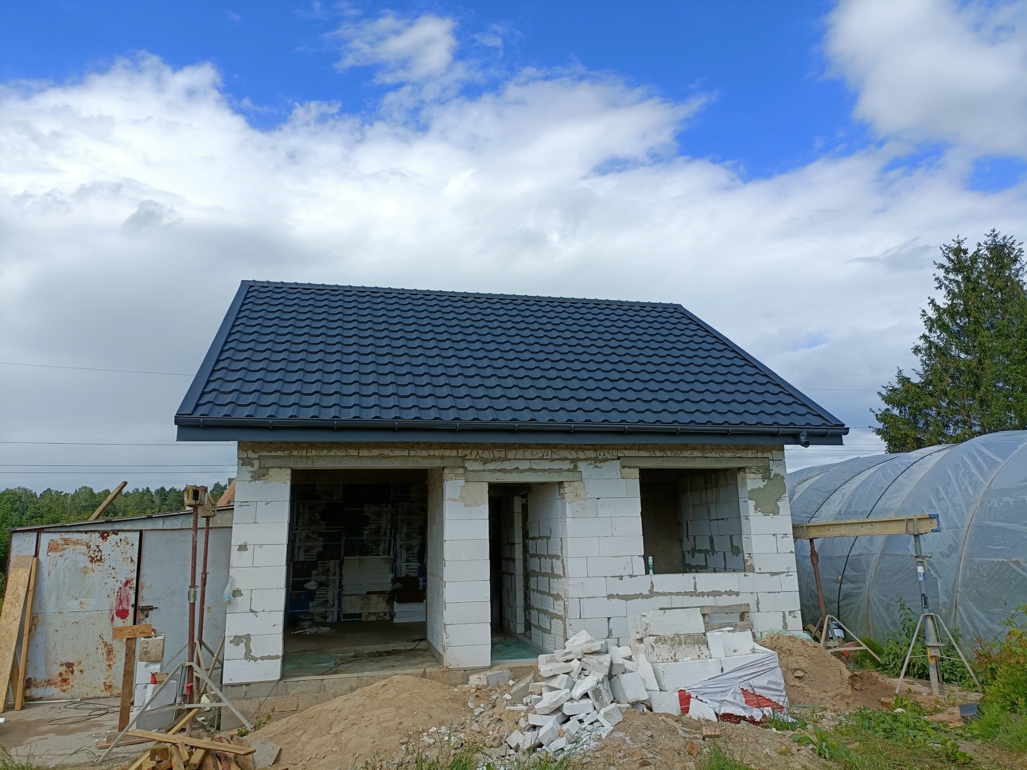 Niedokończony budynek z białych bloczków z ciemnoszarą, metalową dachówką, widoczne otwory okienne i drzwiowe, gruz budowlany na ziemi, w tle szklarnia i błękitne niebo z chmurami.