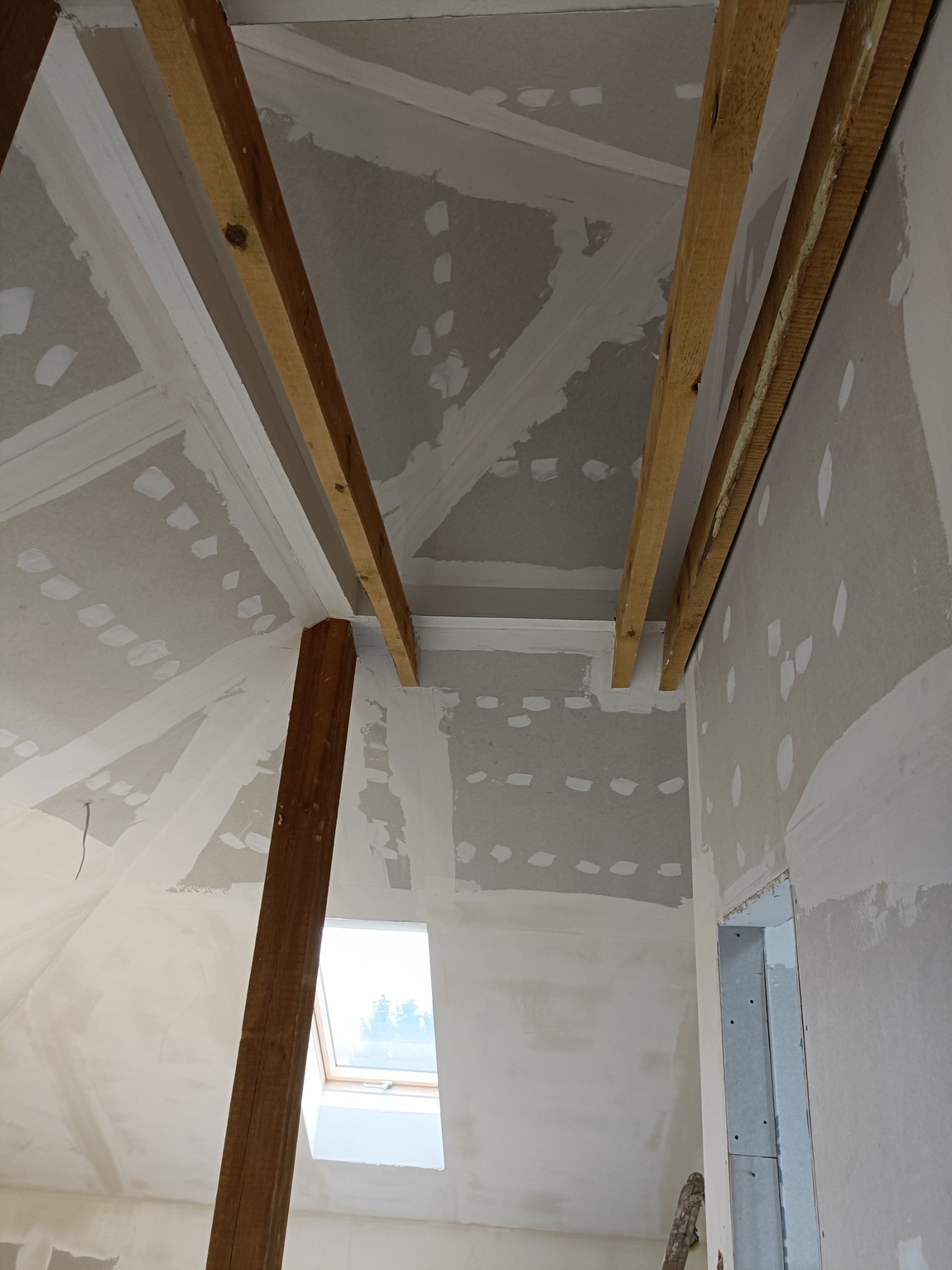Poddasze w trakcie wykańczania, widoczne drewniane belki konstrukcyjne, płyty gipsowo-kartonowe z widocznymi łączeniami przed szpachlowaniem, okno dachowe w skosie, ściany przygotowane pod malowanie.