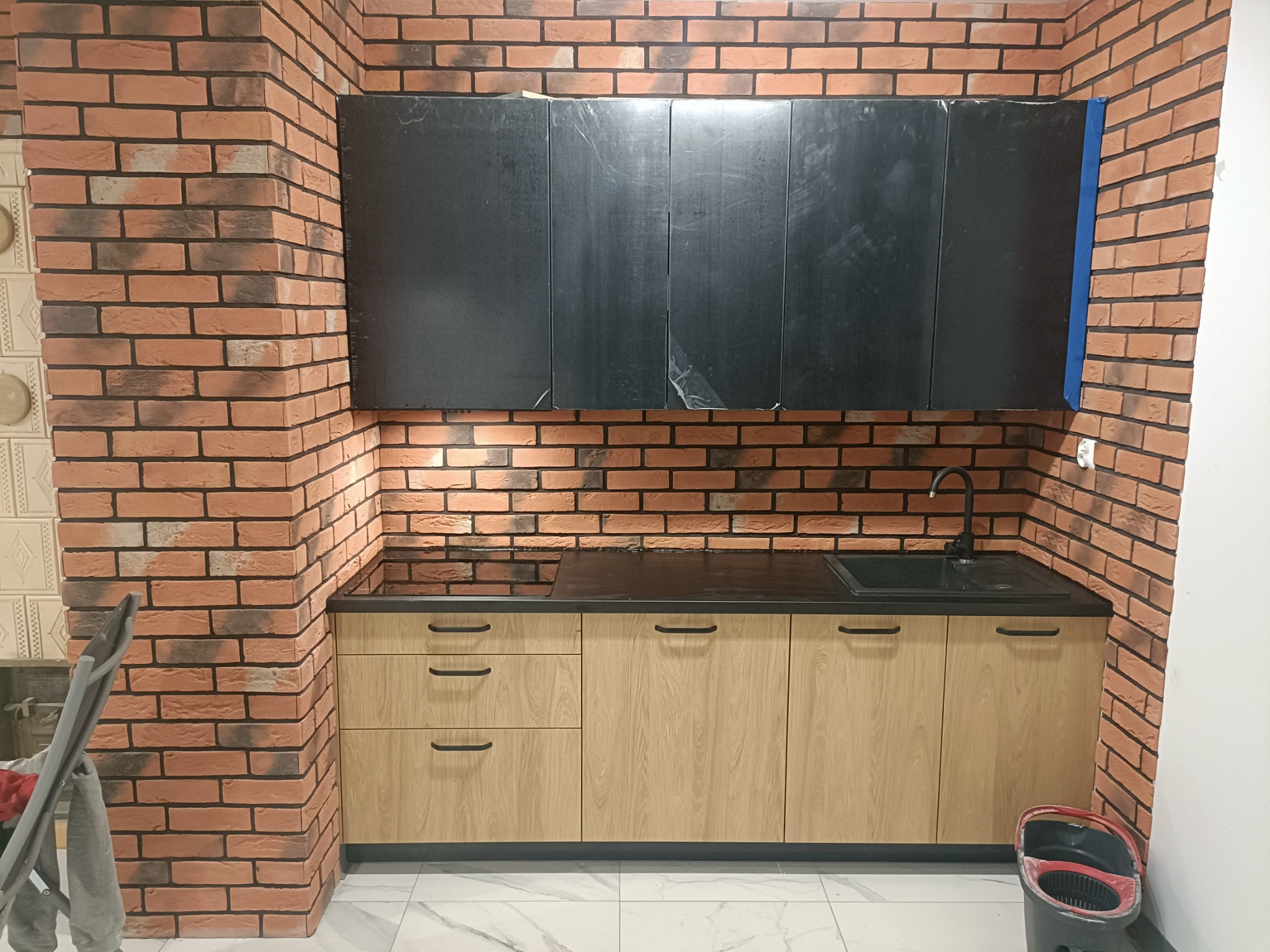 Nowoczesna kuchnia z ceglaną ścianą, drewnianymi szafkami i czarnym blatem oraz umywalką, górne szafki w kolorze grafitowym, widoczny mop w wiadrze.