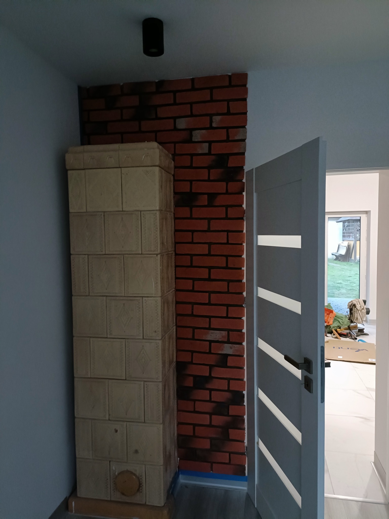Kafelkowy piec kaflowy obok ściany z czerwonej cegły i otwartych, szarych drzwi z białymi paskami w nowym wnętrzu.