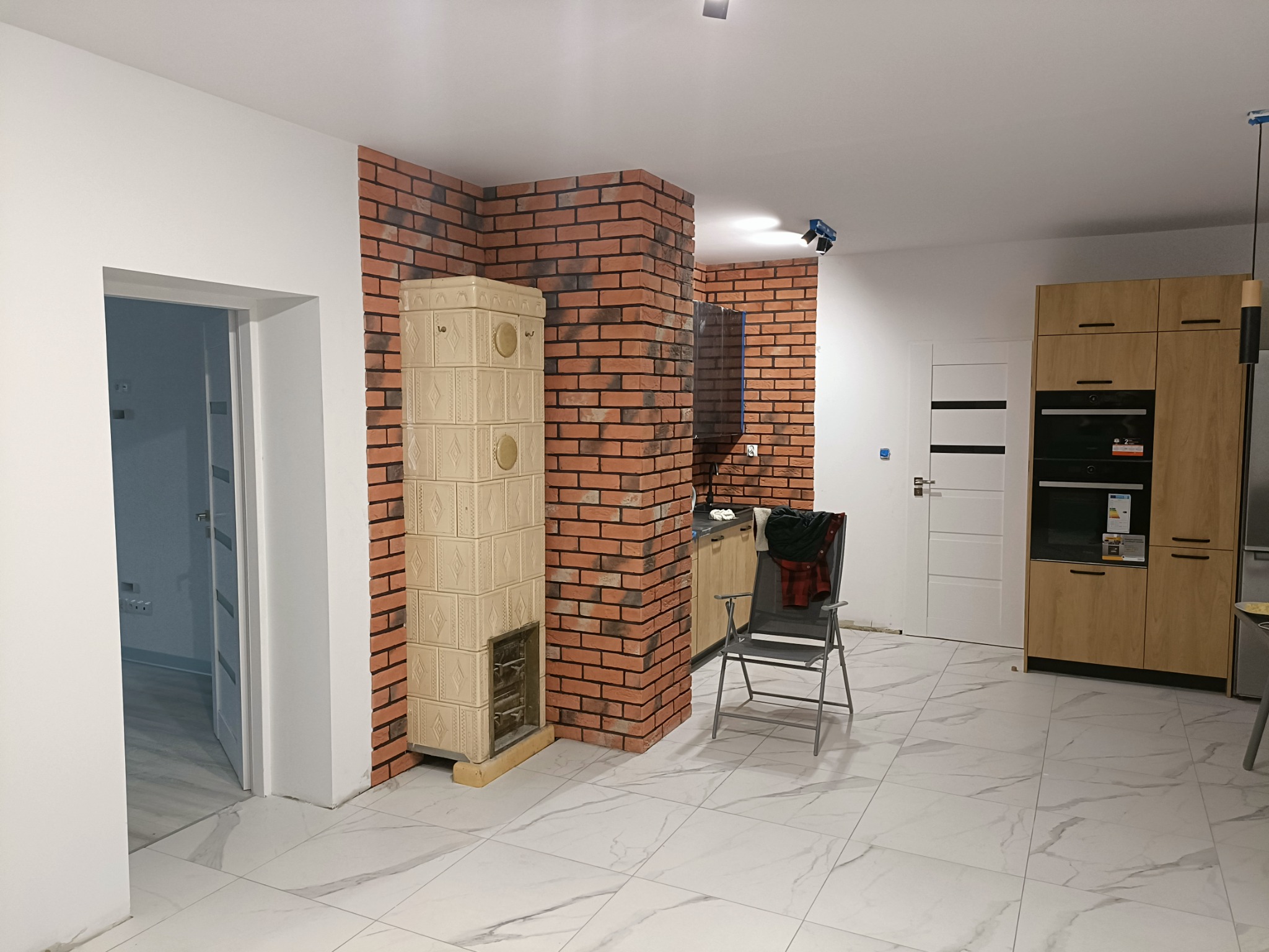 Wykończone wnętrze z ceglaną ścianą, kaflowym piecem i marmurową podłogą, widok na kuchnię z drewnianymi szafkami i metalowym krzesłem.