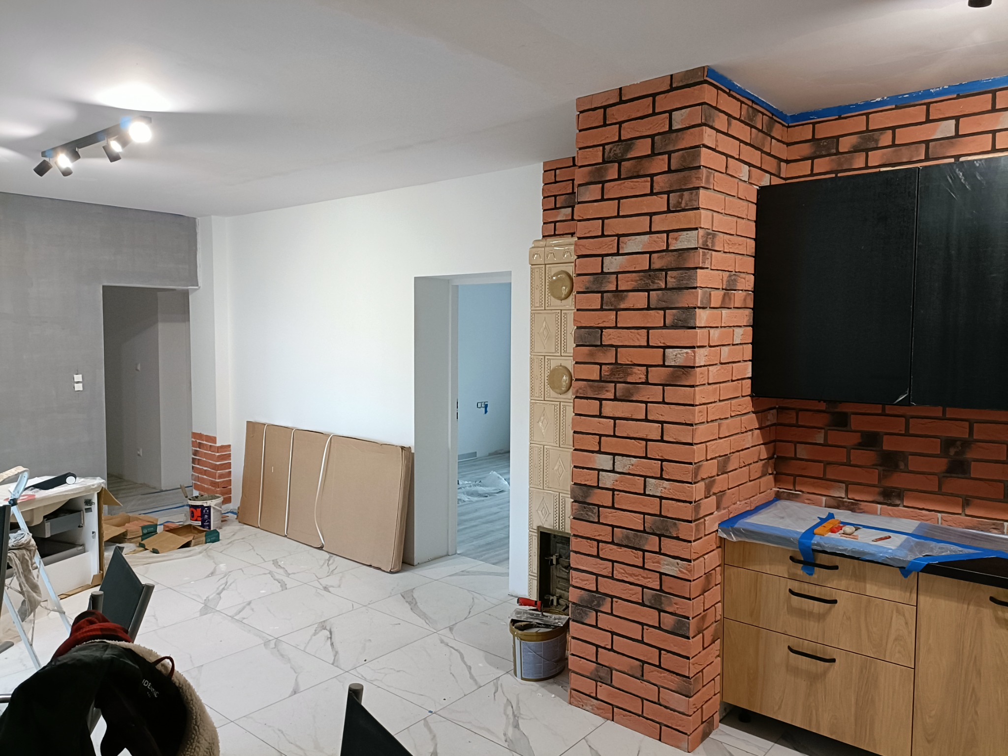 Wnętrze w trakcie prac wykończeniowych: ułożona cegła dekoracyjna na ścianie, zabezpieczone folią meble kuchenne, marmurowe płytki podłogowe, ściany częściowo pomalowane na biało, w tle widoczne...