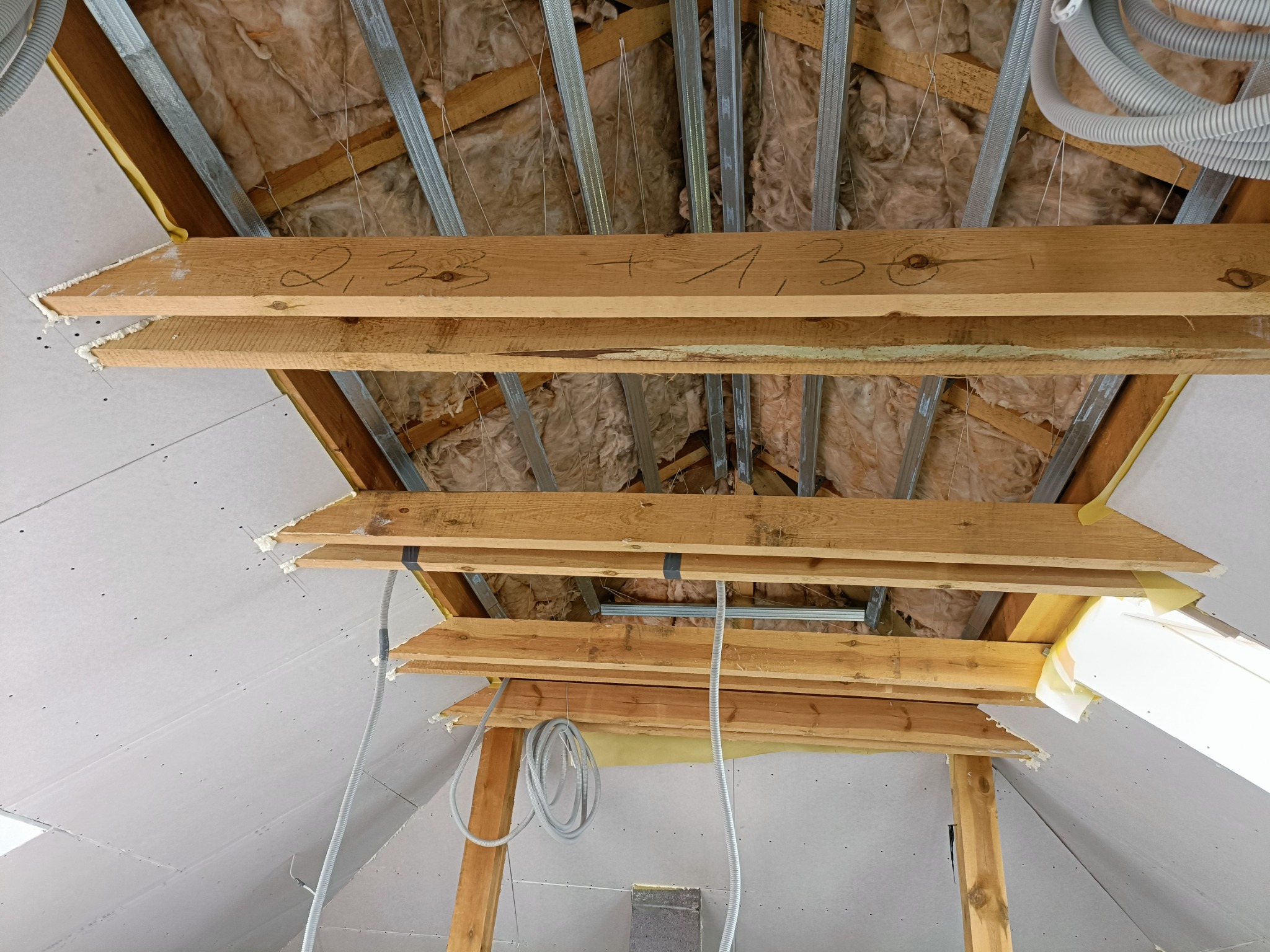 Poddasze w trakcie prac: widoczne ocieplenie wełną mineralną, metalowe profile, drewniane elementy konstrukcyjne z naniesionymi oznaczeniami, częściowo zamontowane płyty gipsowo-kartonowe...