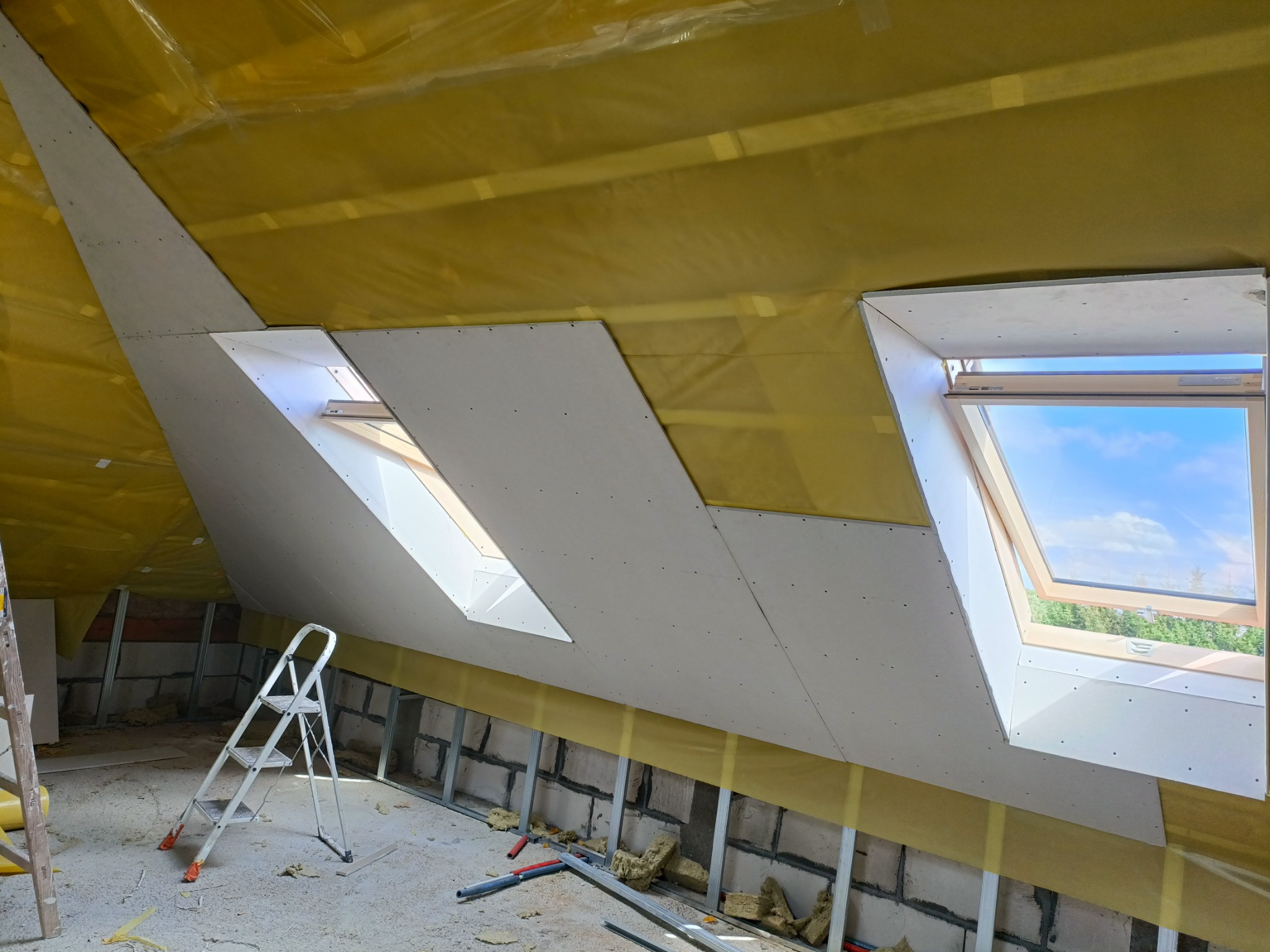 Poddasze w trakcie adaptacji: częściowo zamontowane płyty gipsowo-kartonowe wokół okien dachowych, widoczna izolacja z folii i drabina.