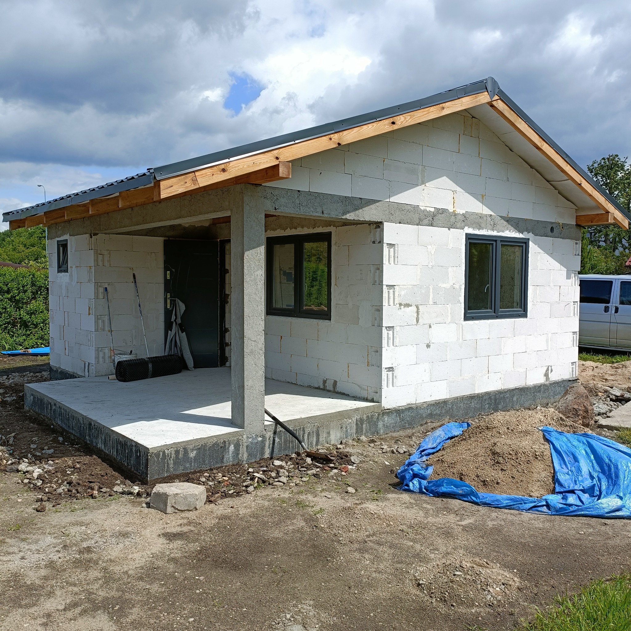 Surowy stan domu z białych bloczków, widoczne szare okna, betonowy fundament i częściowo wykończony dach z drewnianą konstrukcją, na pierwszym planie sterta piasku przykryta niebieską plandeką.