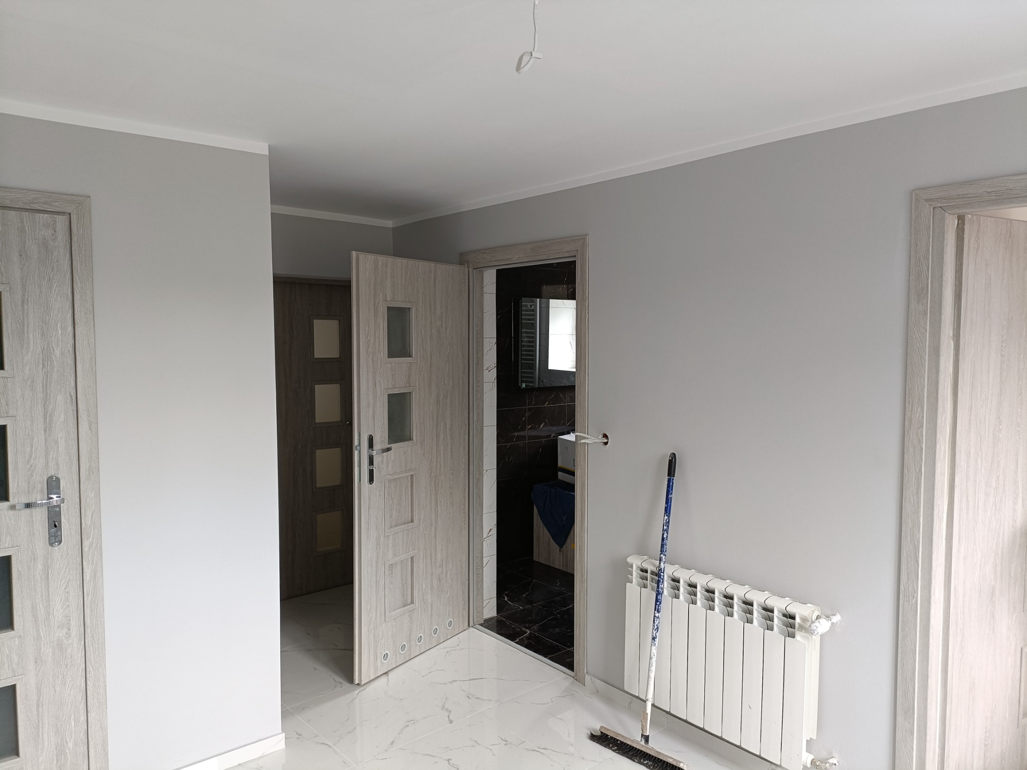 Jasne, szare wnętrze po remoncie z widocznymi, nowymi drzwiami z przeszkleniami, białym grzejnikiem i marmurową podłogą. W tle widoczna łazienka z czarnymi płytkami. Na podłodze stoi szczotka...