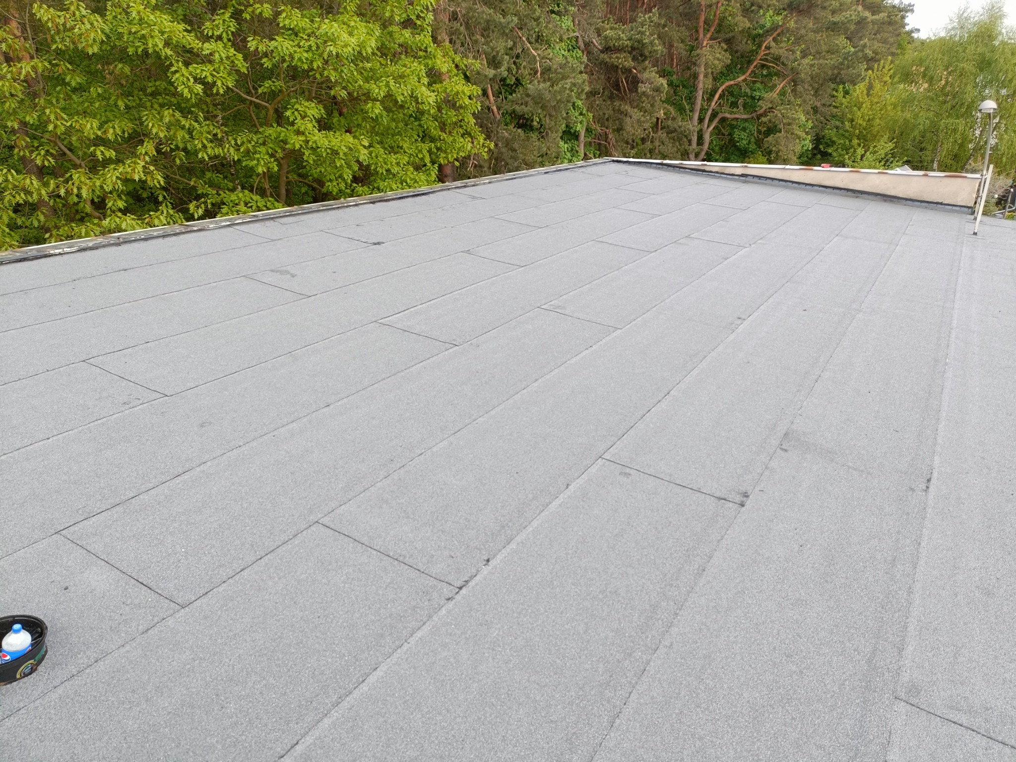 Nowo pokryty, płaski dach z papy termozgrzewalnej w kolorze szarym, widoczne łączenia, w tle zieleń drzew, na dachu wiaderko z wodą.